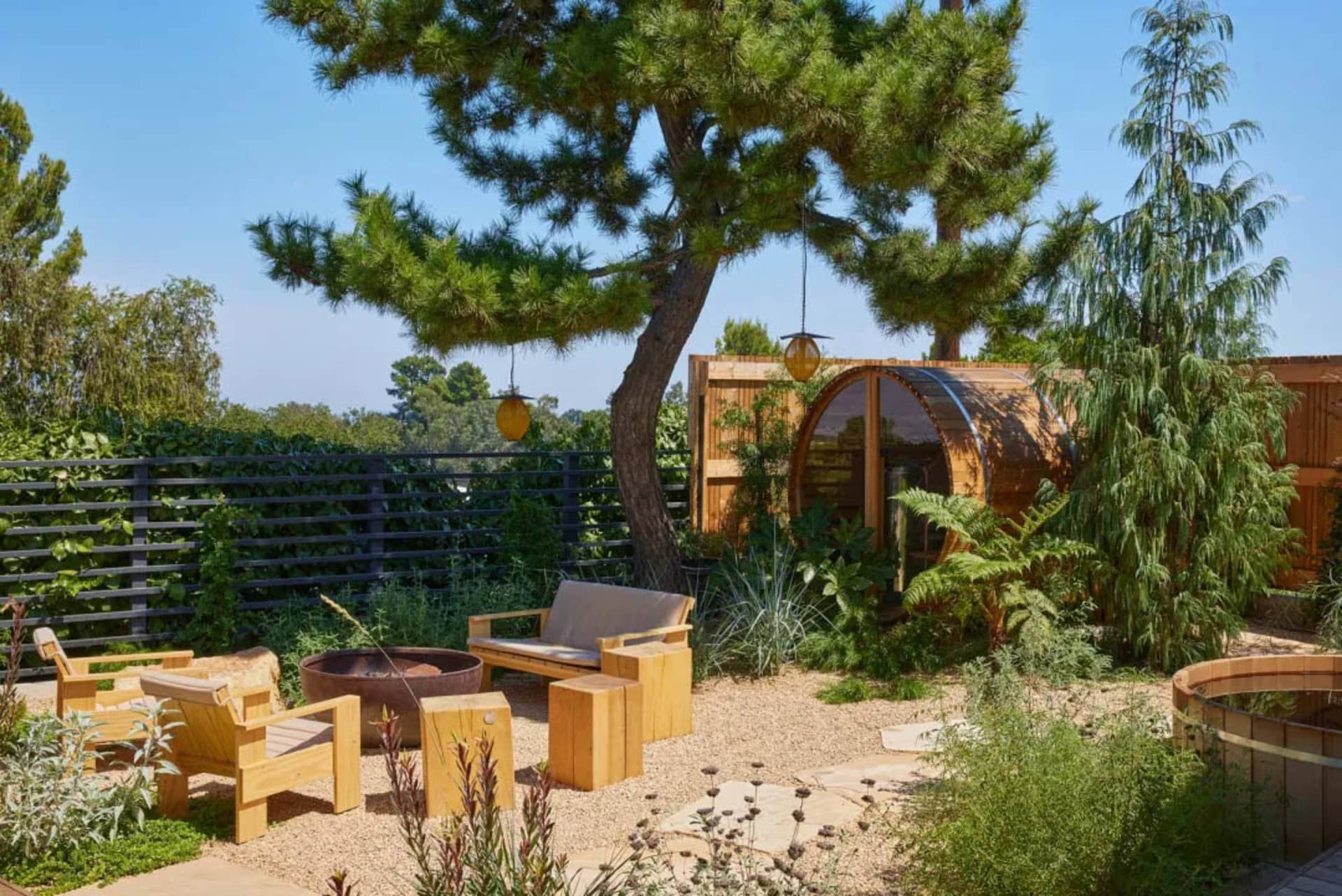 A serene outdoor patio with a wooden deck, gazebo, and natural surroundings, featuring a wooden bench and chair on the dirt ground