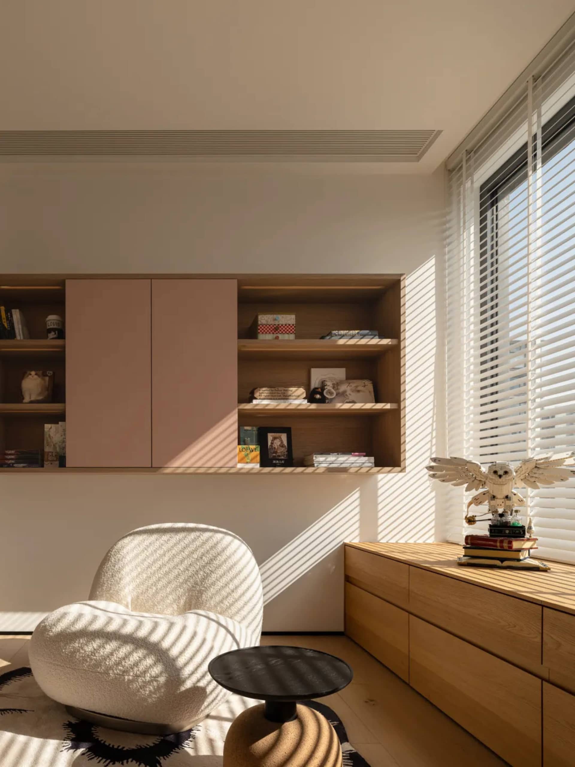 A modern living room with a white chair, a bookshelf, and a window, featuring a clean and minimalist design with white walls and a wooden floor, allowing natural light to enter the room