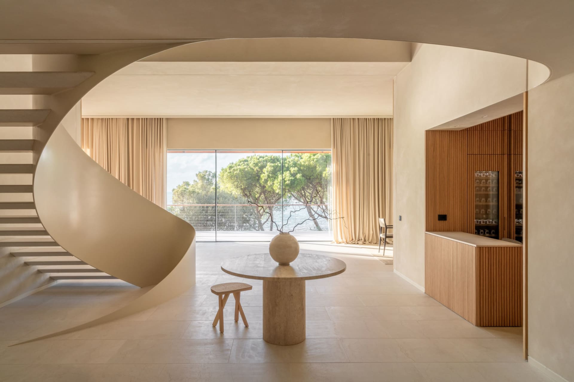 A modern living and dining space with a white color scheme, featuring a large table, chair, couch, and dining table, with natural light streaming in through the windows