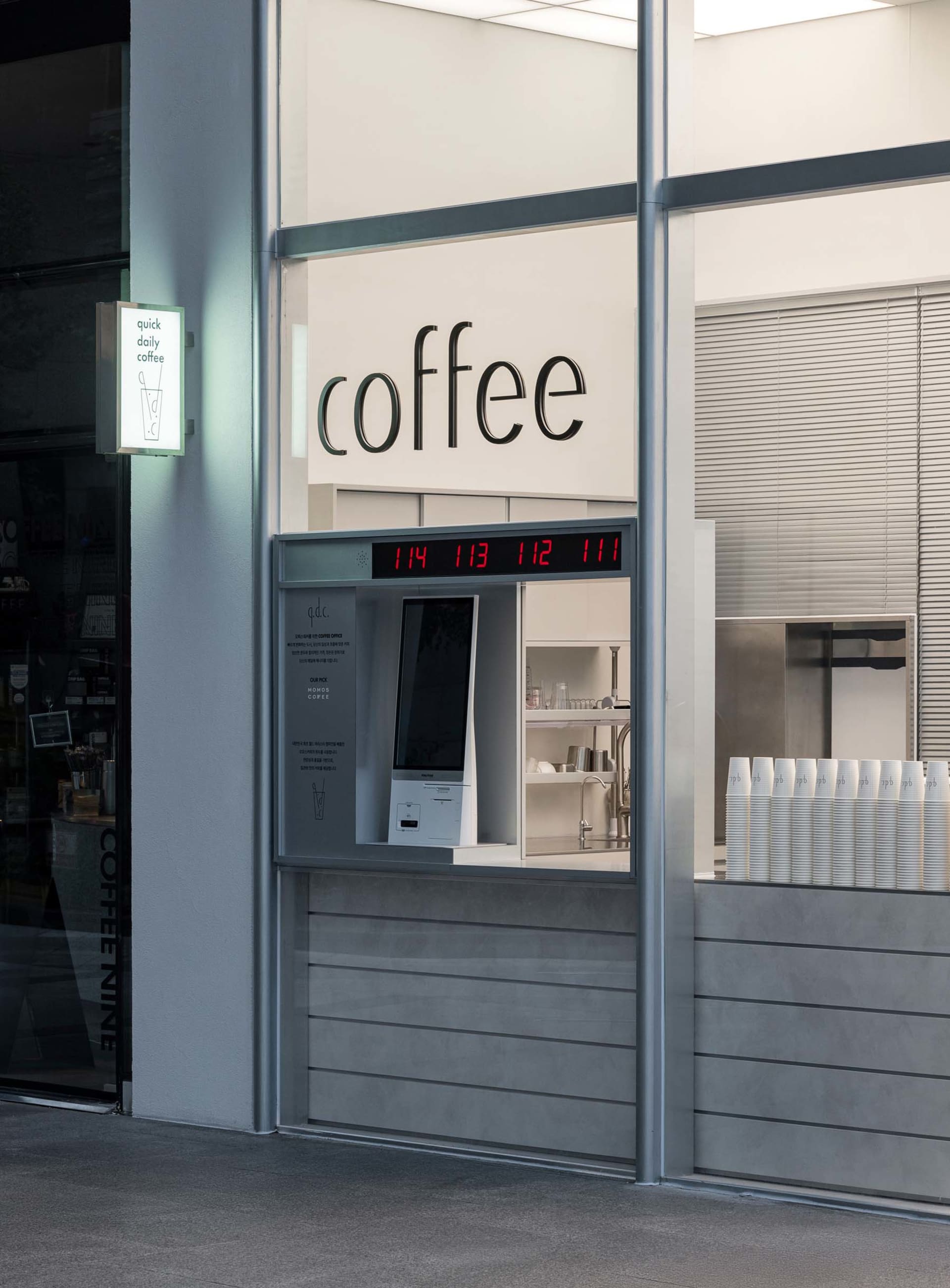 A modern coffee shop with a glass front, white walls, and a sleek metal counter, featuring a coffee machine, cups, and bottles, under bright and welcoming lighting