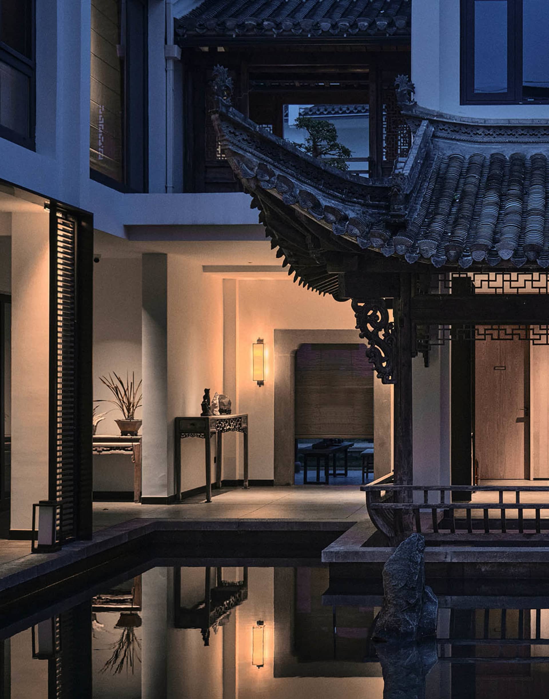 A well-lit room with a Chinese-style roof, featuring a pool, dining table, and bench, decorated with plants, vases, and a bowl, with warm wood and stone materials
