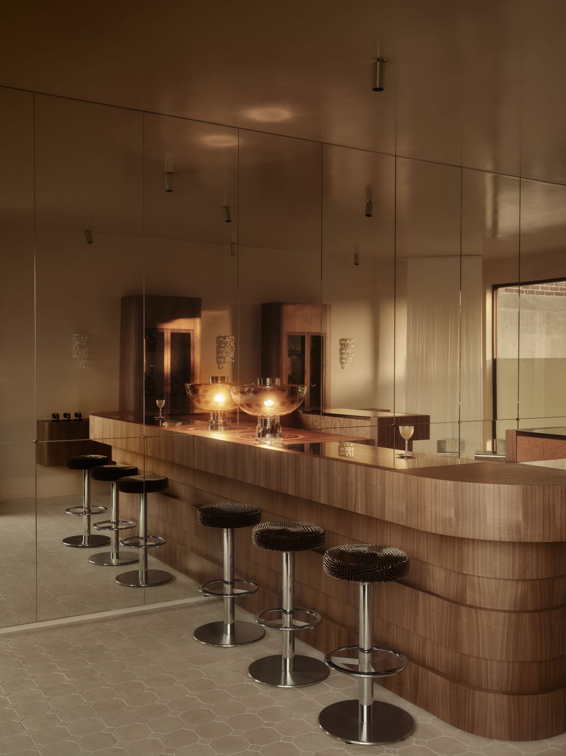 Modern bar area with a wooden counter, large mirror, and glass wall, dimly lit for a cozy atmosphere