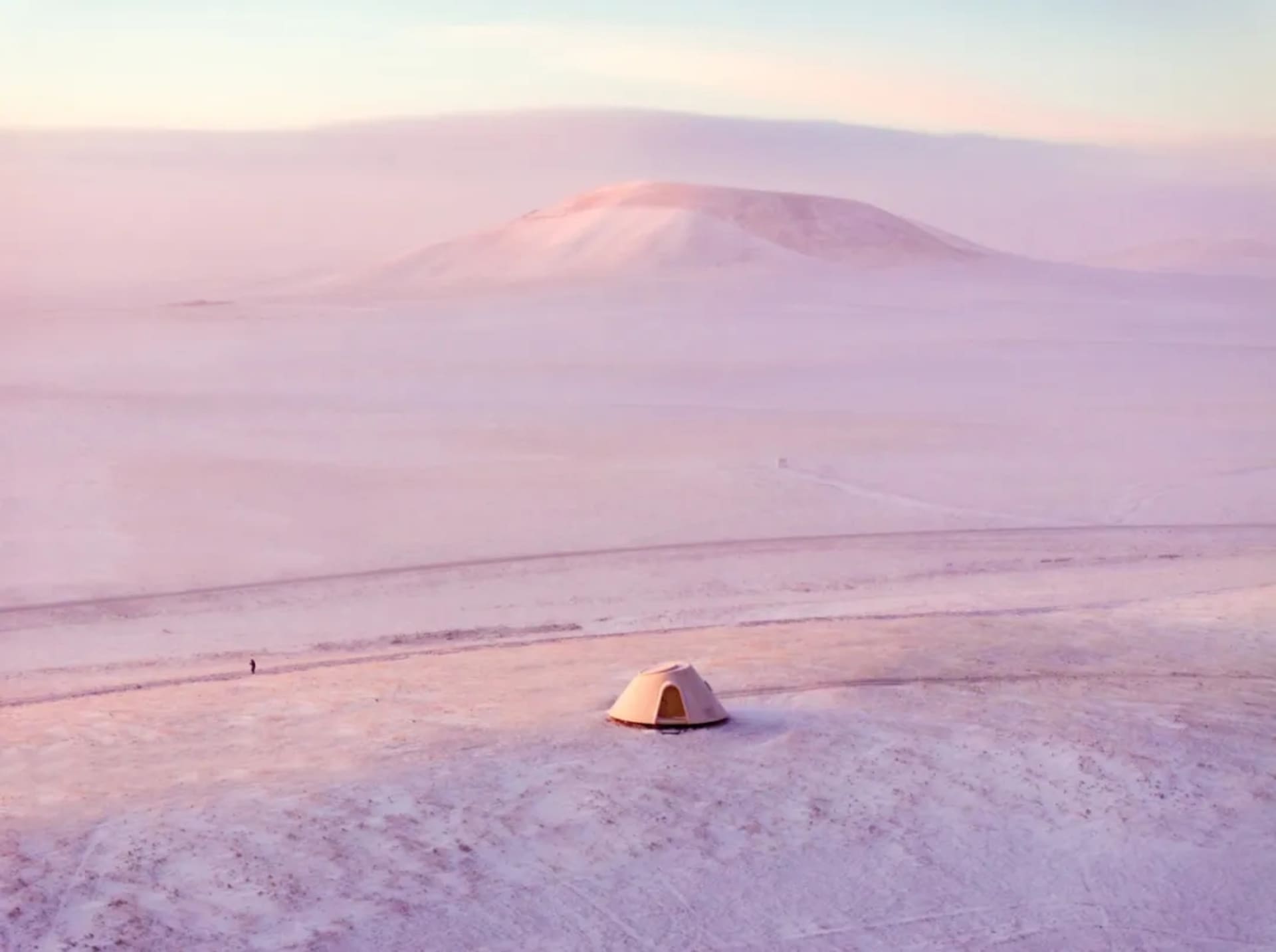 A snowy landscape with a tent in the middle, surrounded by a field and a person standing nearby