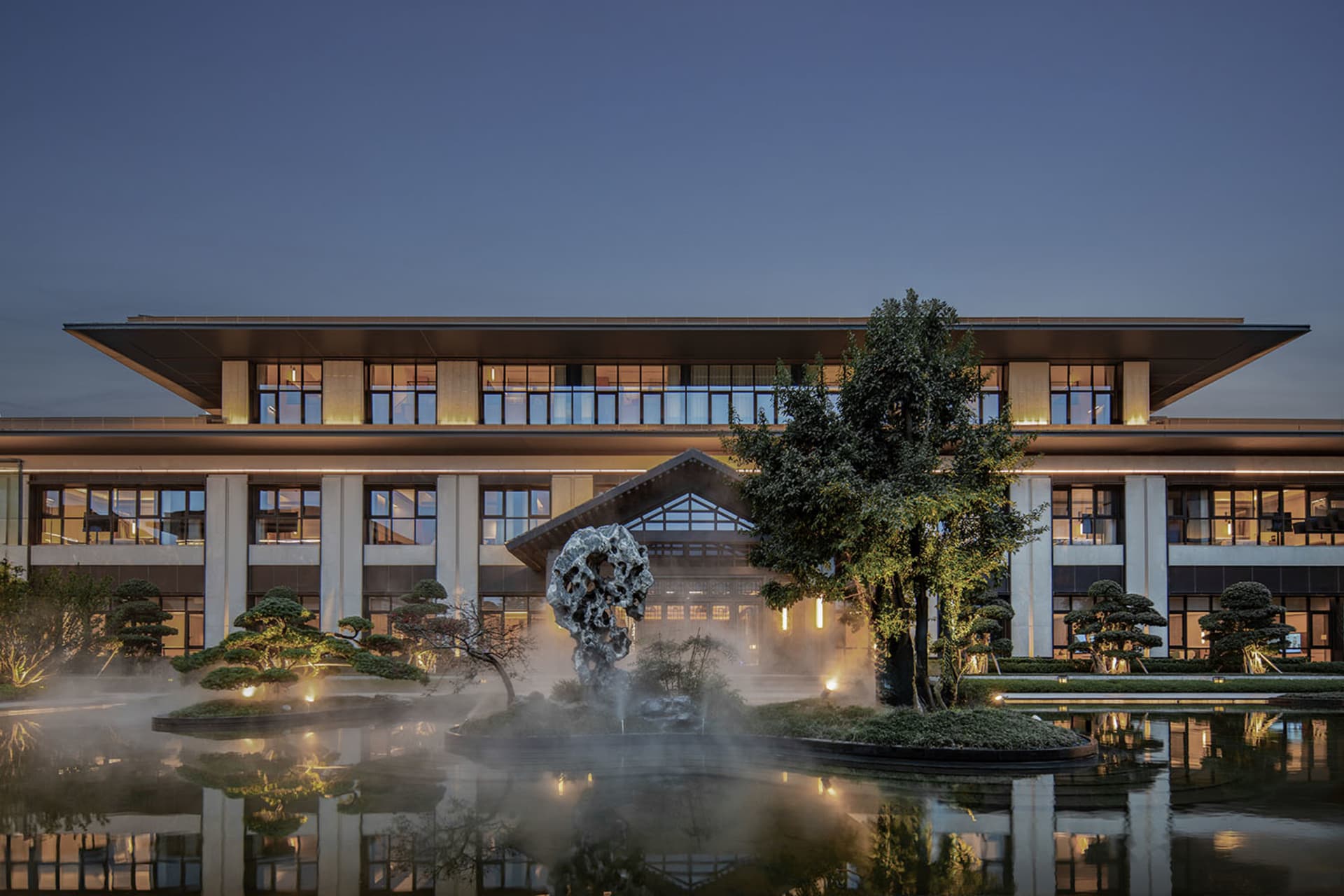 A serene modern building with a glass facade and a pond surrounded by trees, set against a soft and diffused light background