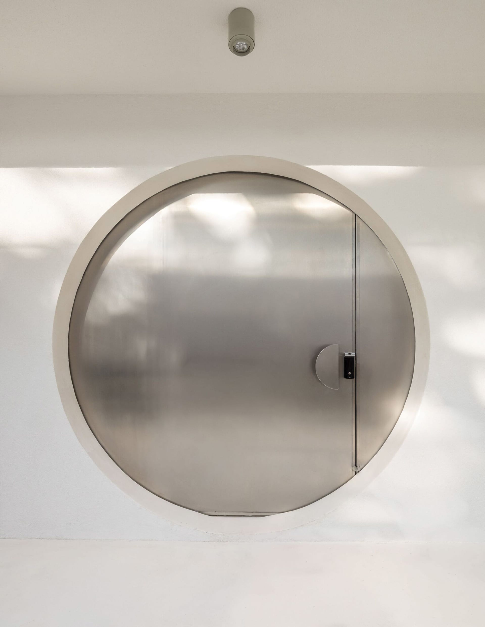 A modern doorway with a circular metal door in a white hallway, lit by natural daylight