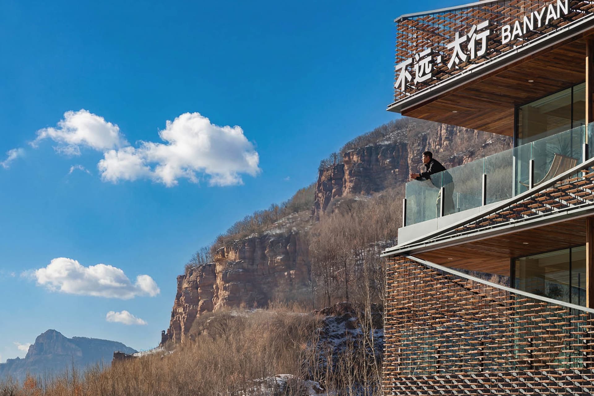A man stands on a glass balcony overlooking a mountainous landscape with a clear blue sky and natural lighting