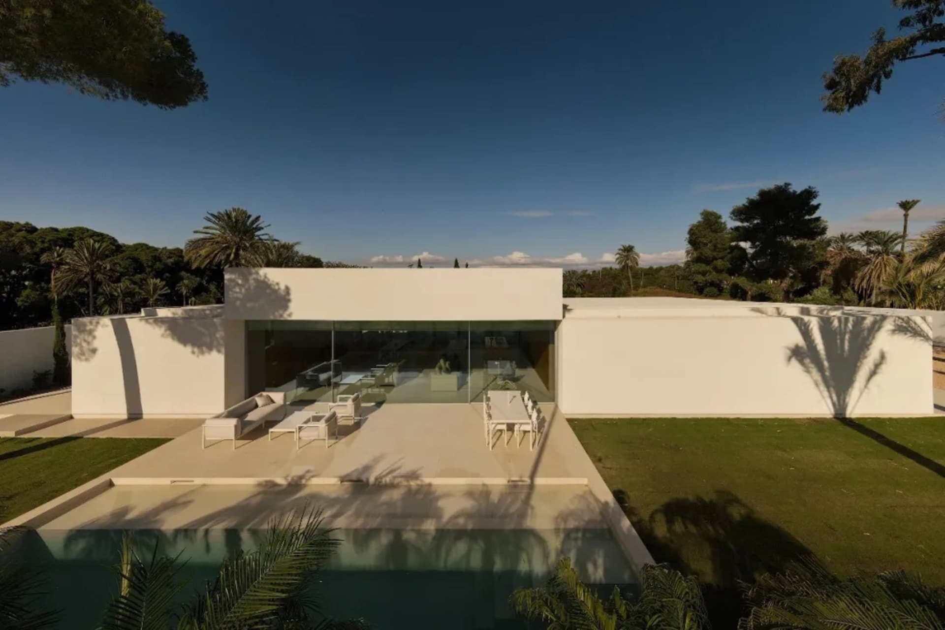 A modern house with a large glass wall, pool, and patio, surrounded by white materials and natural light