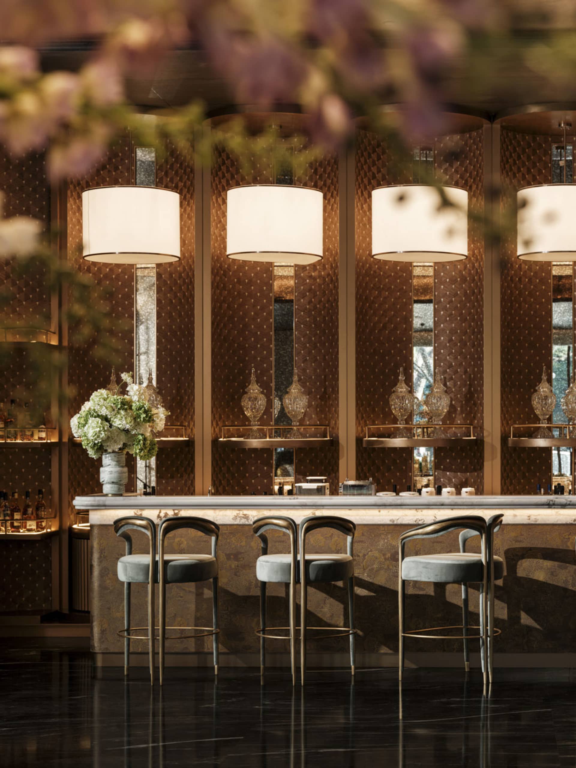 A modern bar with a marble counter, stools, and a vase of flowers, decorated with bottles and cups, and two potted plants, under soft and warm lighting