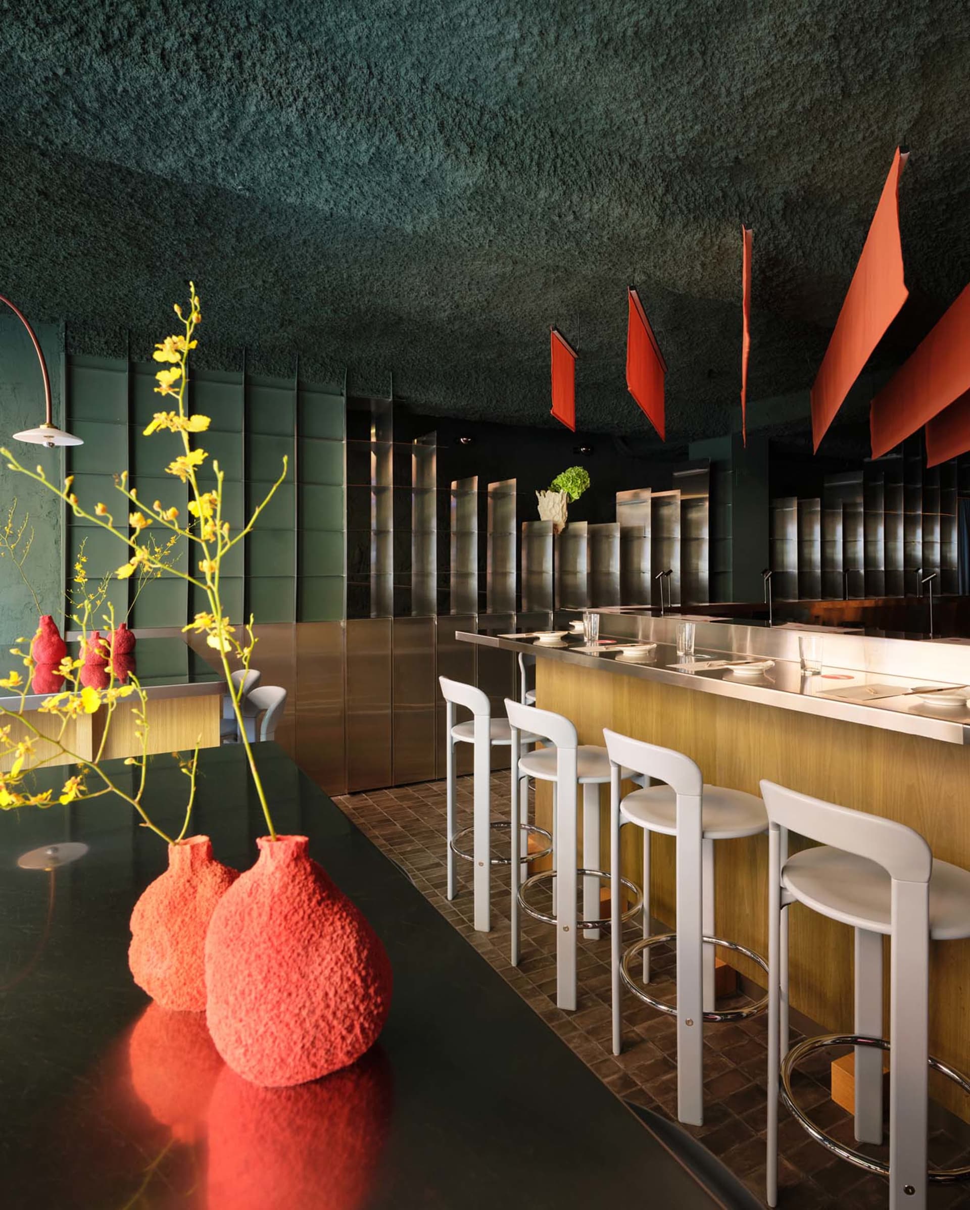 A modern kitchen with a black counter, white stools, and a red vase, lit by natural light