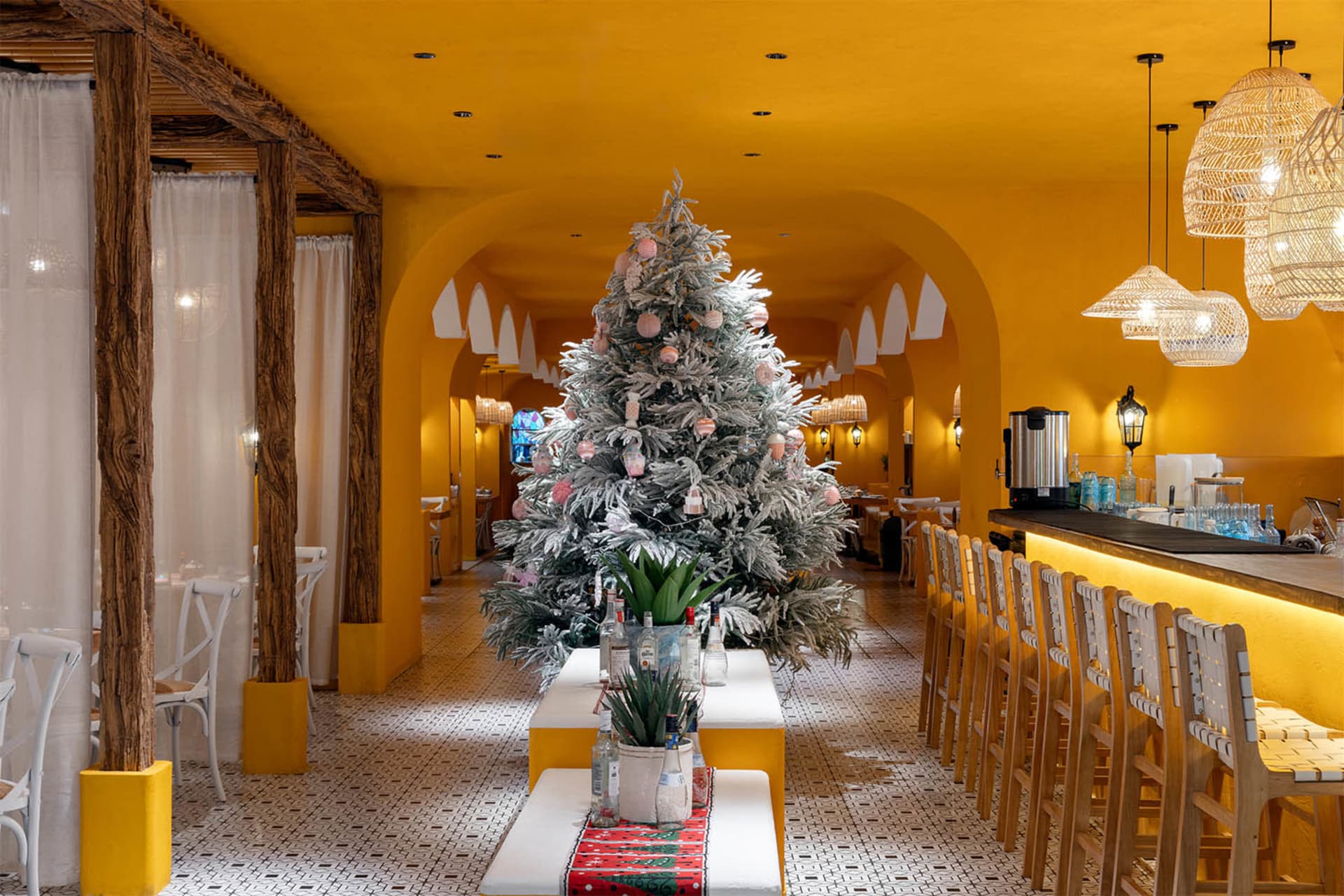 A cheerful dining area with a large Christmas tree, decorated with ornaments, surrounded by chairs and tables in a warm yellow-painted room