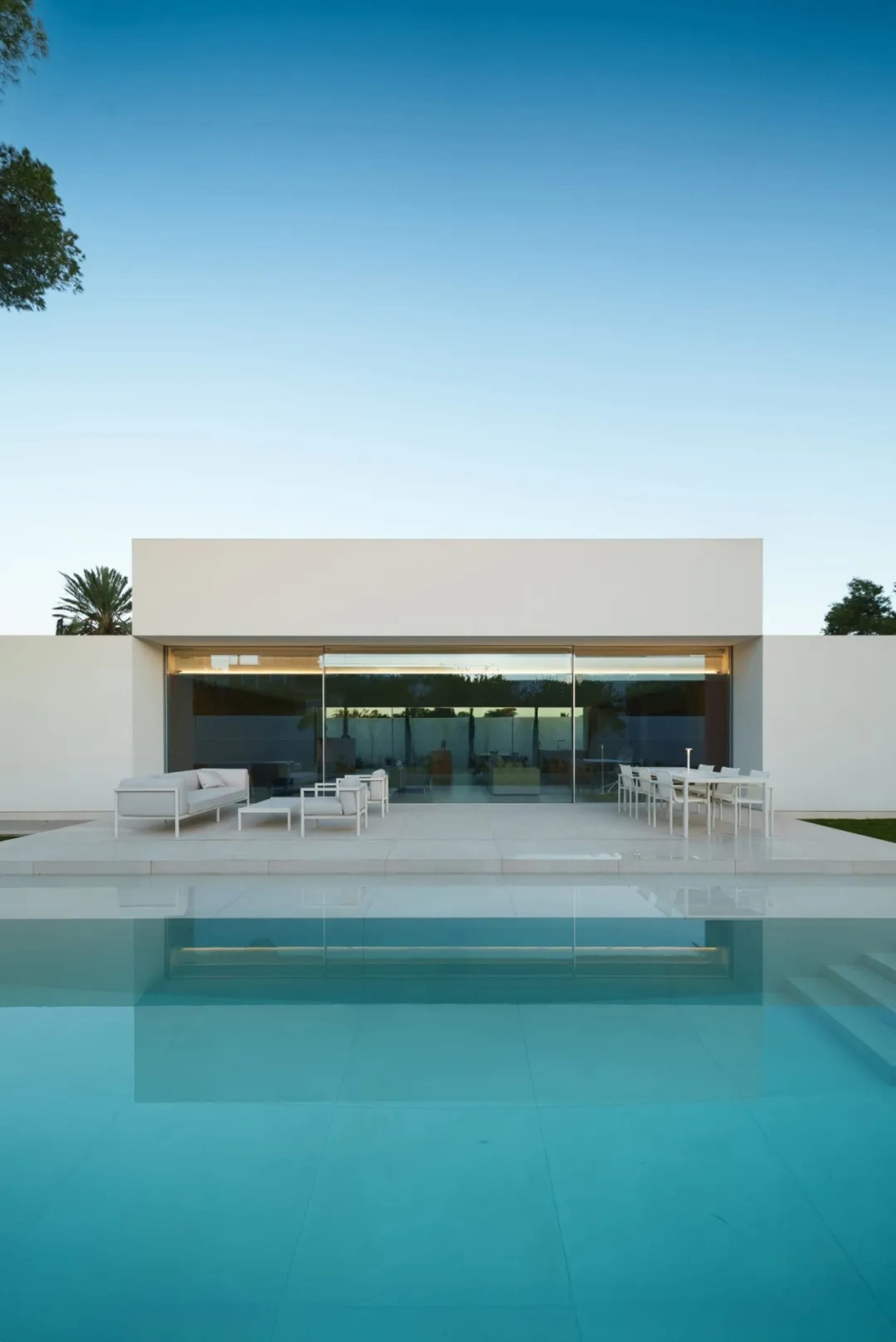 A modern house with a large glass wall, pool, and patio area, surrounded by white materials and natural light