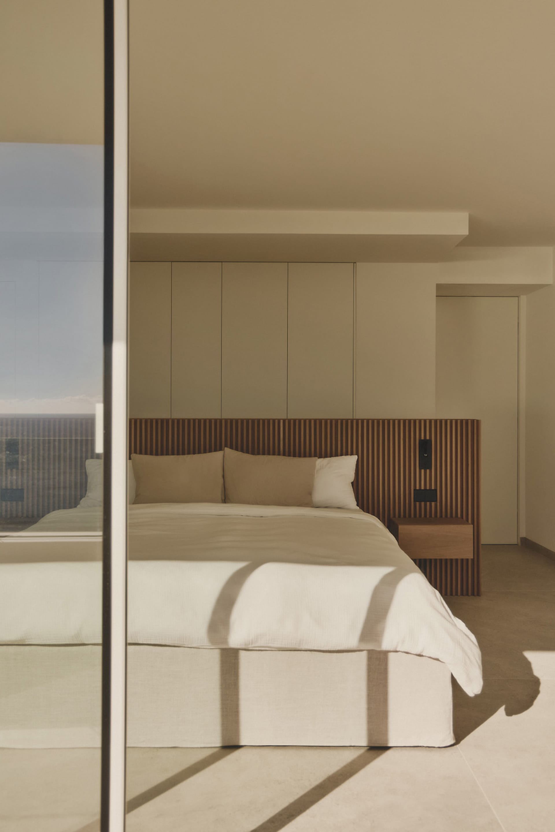 A well-lit bedroom with a large bed, a dresser, and a window, featuring predominantly white materials and a clean, modern design
