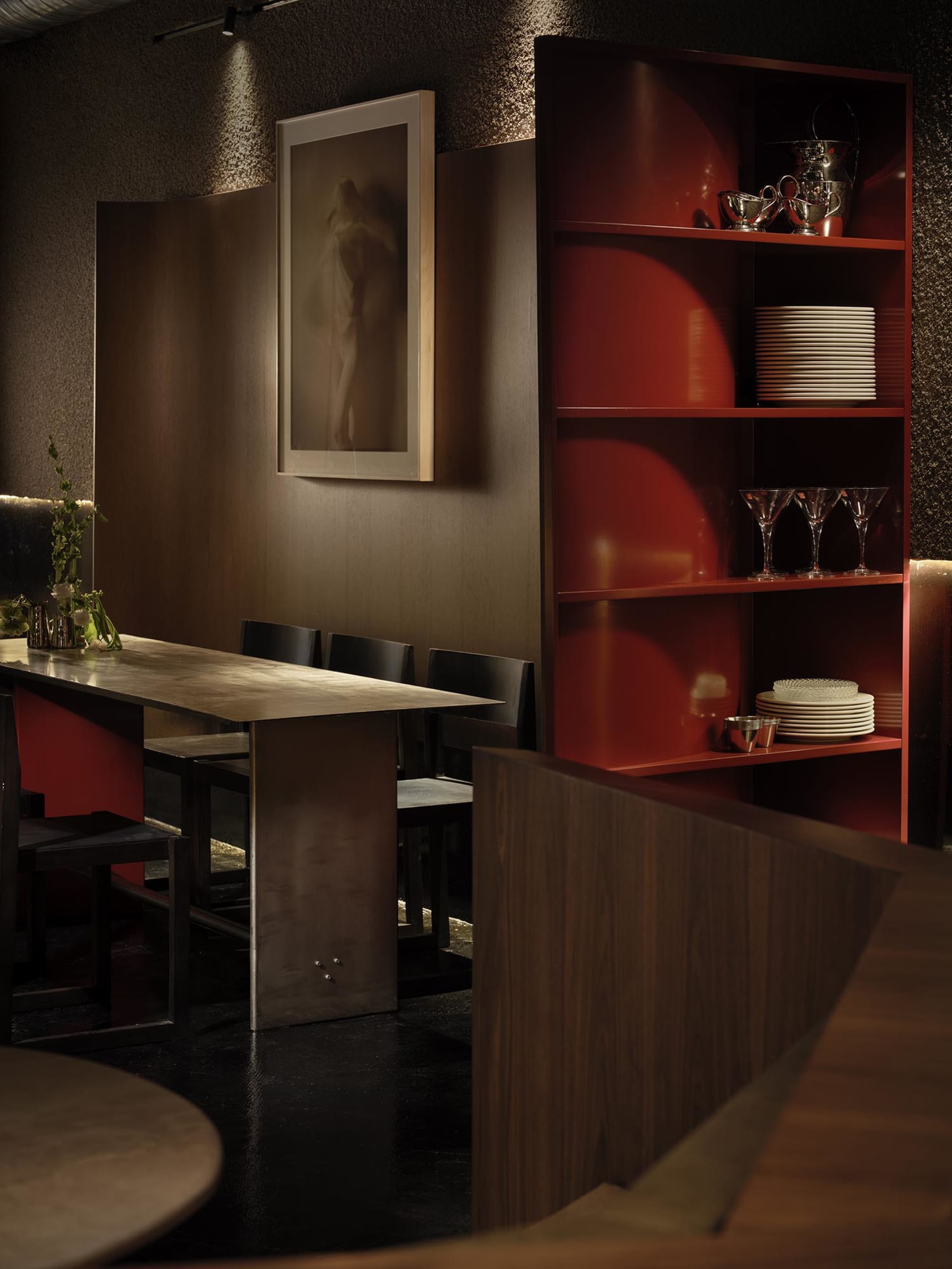 A modern dining room with a wooden table and chairs, surrounded by a red bookshelf, in a dimly lit space