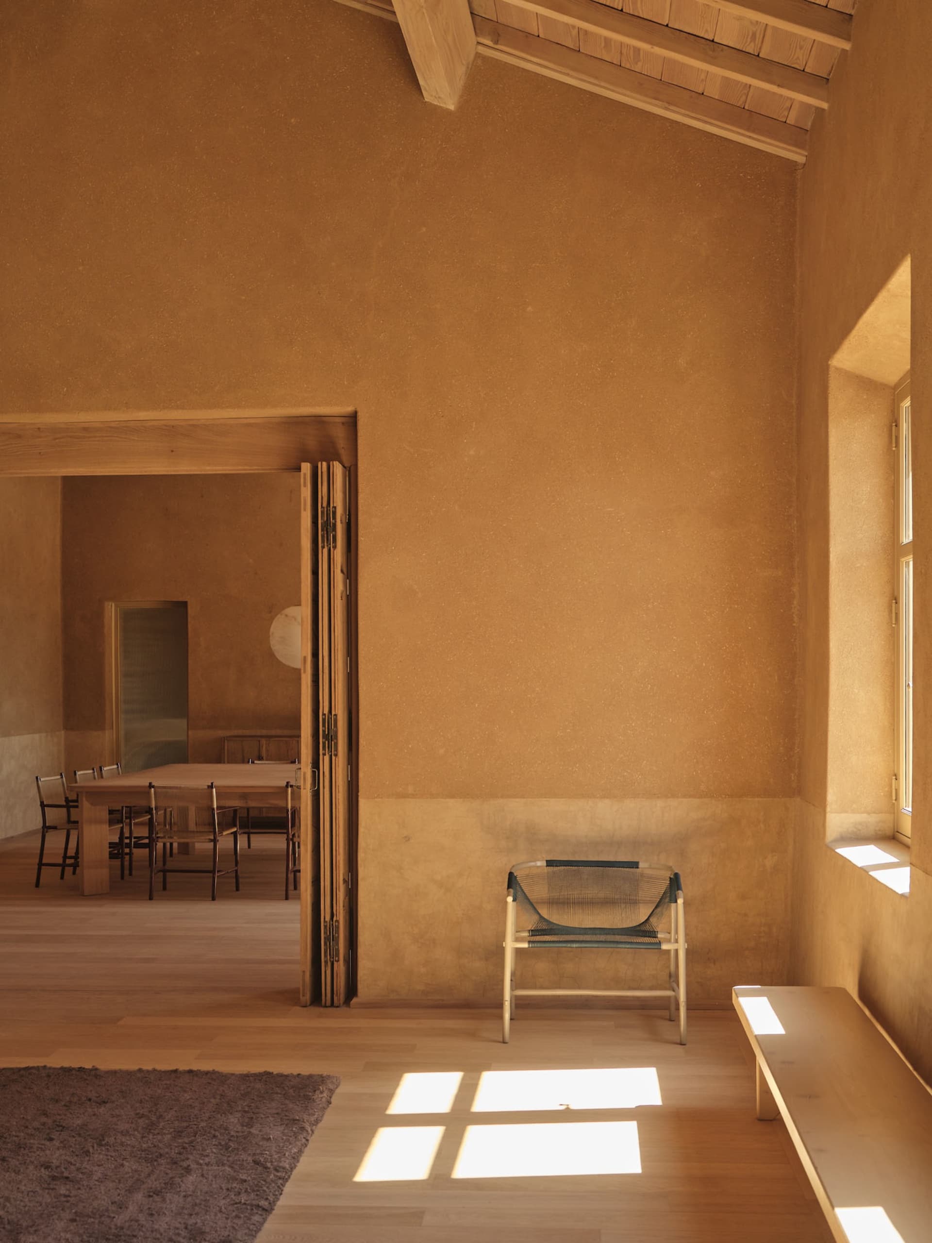 Cozy dining space with a wooden floor, a dining table, chairs, and a bench, surrounded by cement walls and natural light from a window