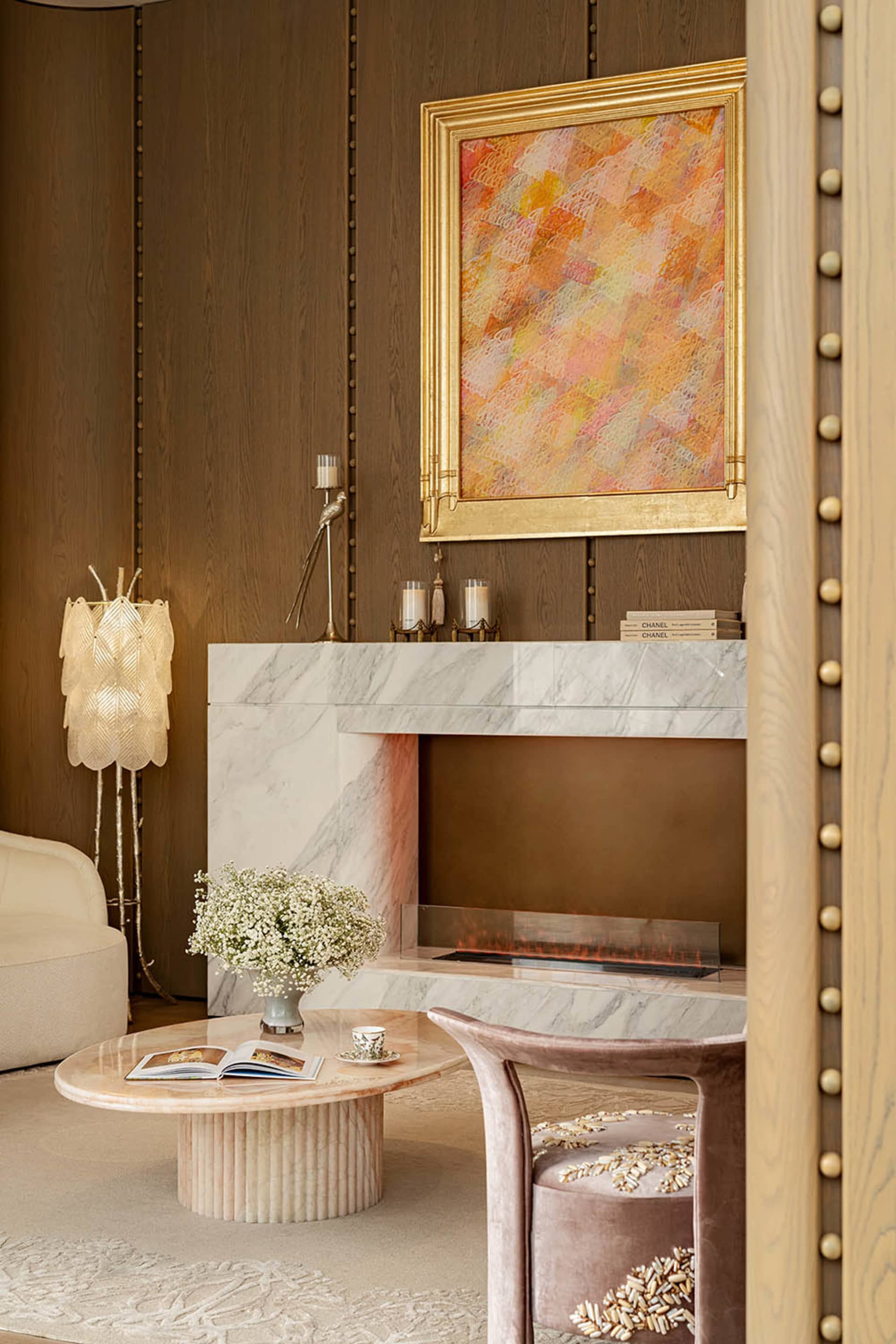 Cozy living room with a pink and white color scheme, featuring a marble fireplace, a couch, and a chair, with a dining table and vase adding to the aesthetic
