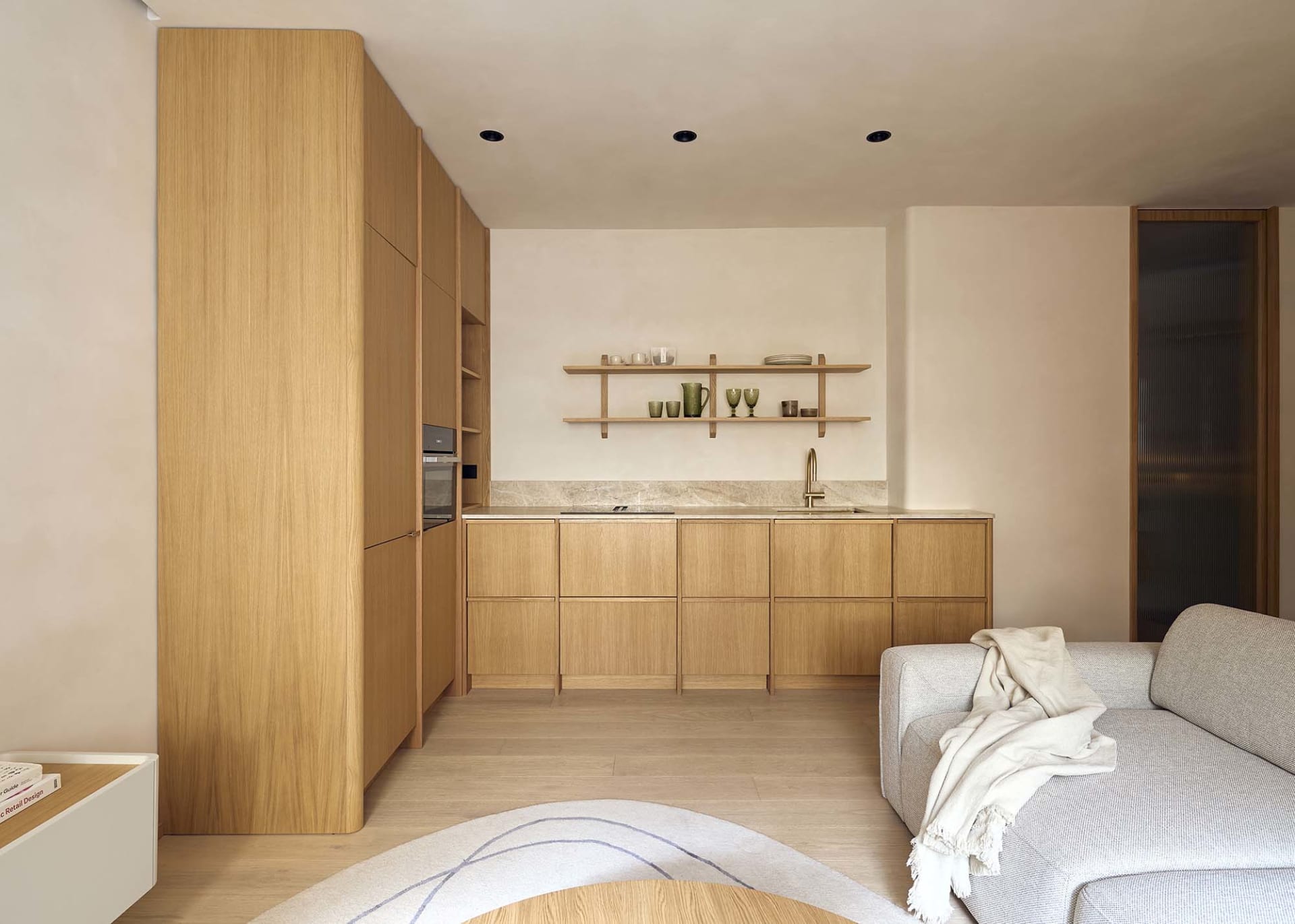 Modern kitchen and living room area with wooden cabinets, a couch, chair, vase, bowl, and bottle, featuring soft and warm lighting