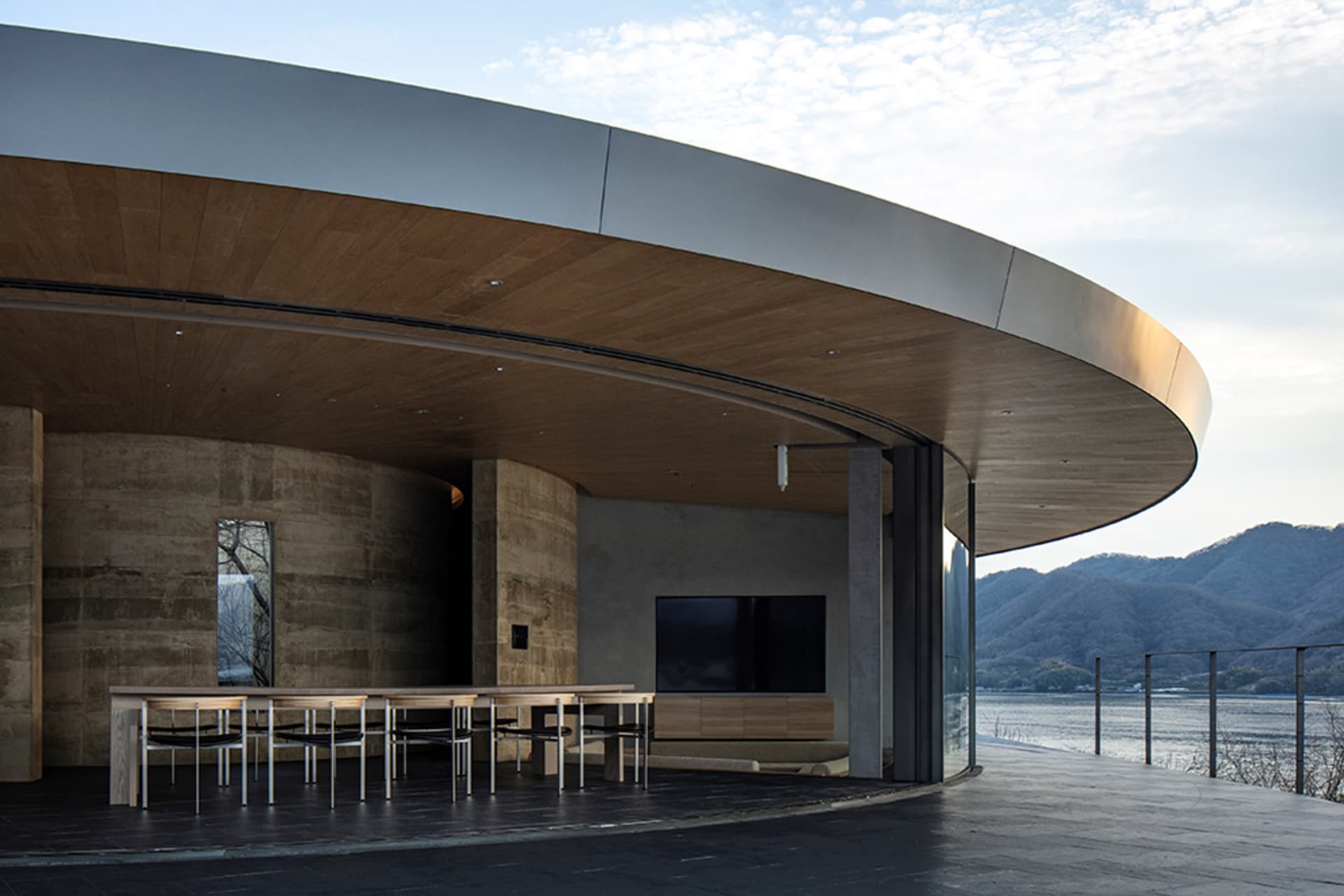 A modern restaurant with a circular seating area featuring a large TV, chairs, and dining tables, surrounded by contemporary materials and well-designed lighting