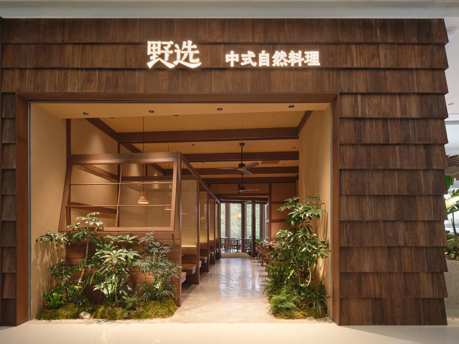 A modern restaurant with a wooden exterior, well-lit interior, and natural materials, featuring furniture, a potted plant, and a welcoming atmosphere