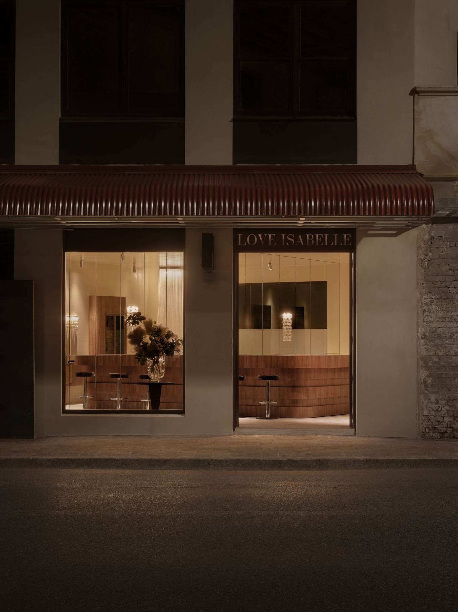 A storefront with a sign reading Love Isabelle, featuring a display of potted plants and chairs in front of a large window, with a warm and inviting atmosphere