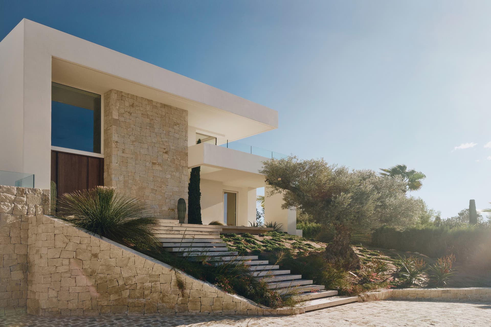 A modern house with a white exterior and a large patio area surrounded by a stone wall, featuring a large glass door and a sunny view