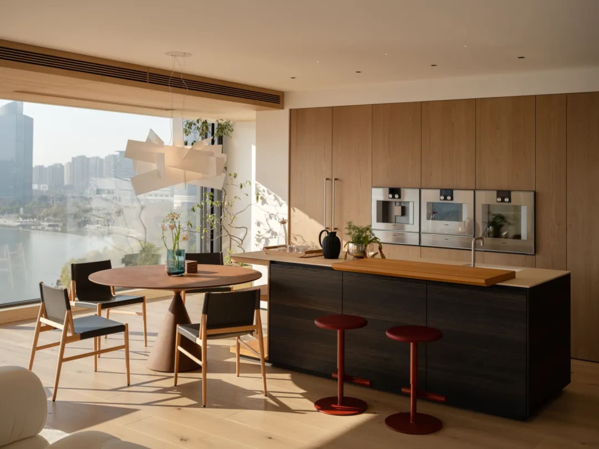 Modern kitchen with a large window, wooden floors, and a dining table, featuring a sink, oven, and refrigerator, surrounded by chairs and potted plants