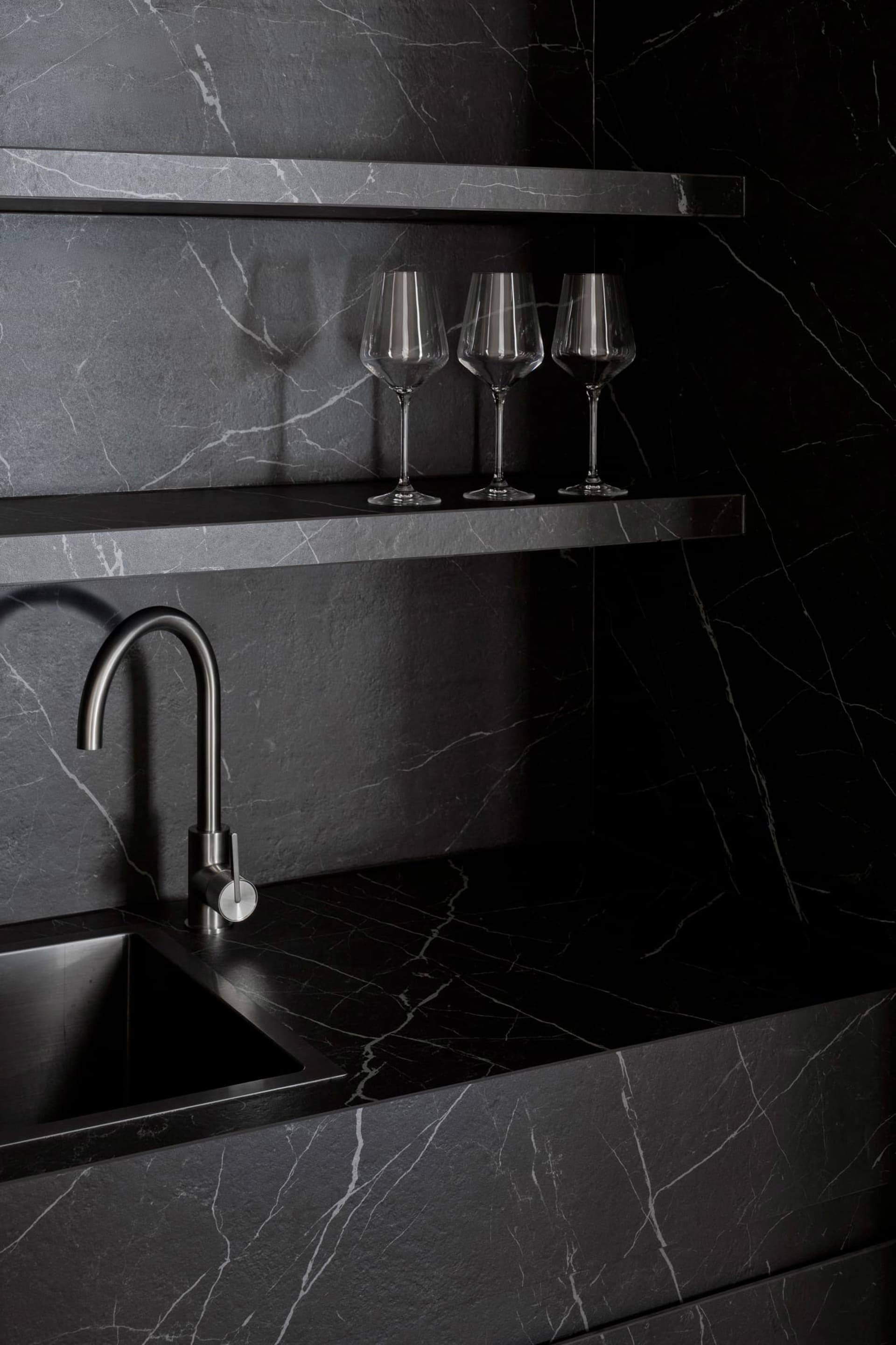 A modern kitchen with a marble counter, sink, and three wine glasses, set in a dimly lit space