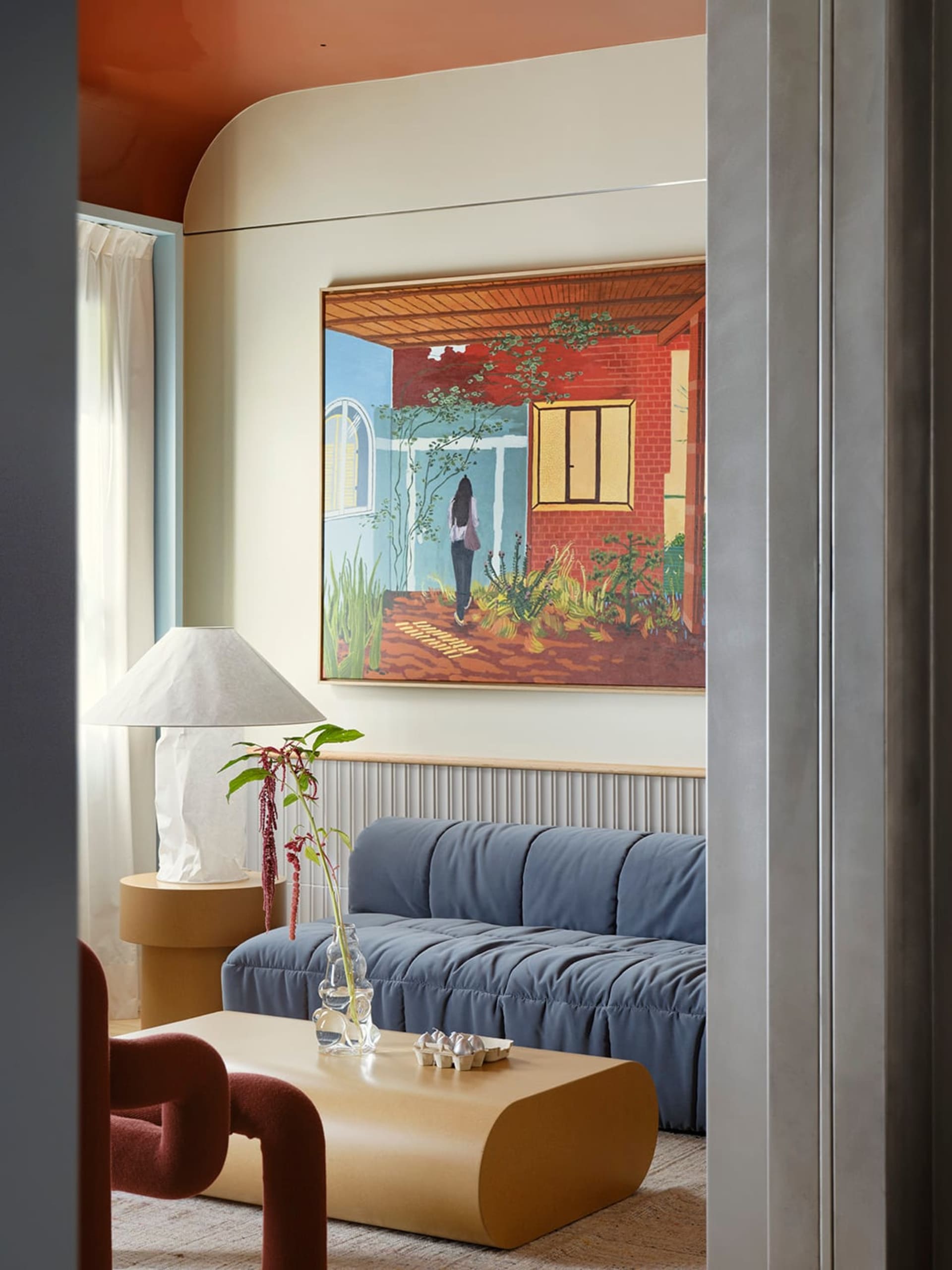Comfortable living space with a blue couch, wooden furniture, and a glass table, featuring a painting and a potted plant, with natural light from a window