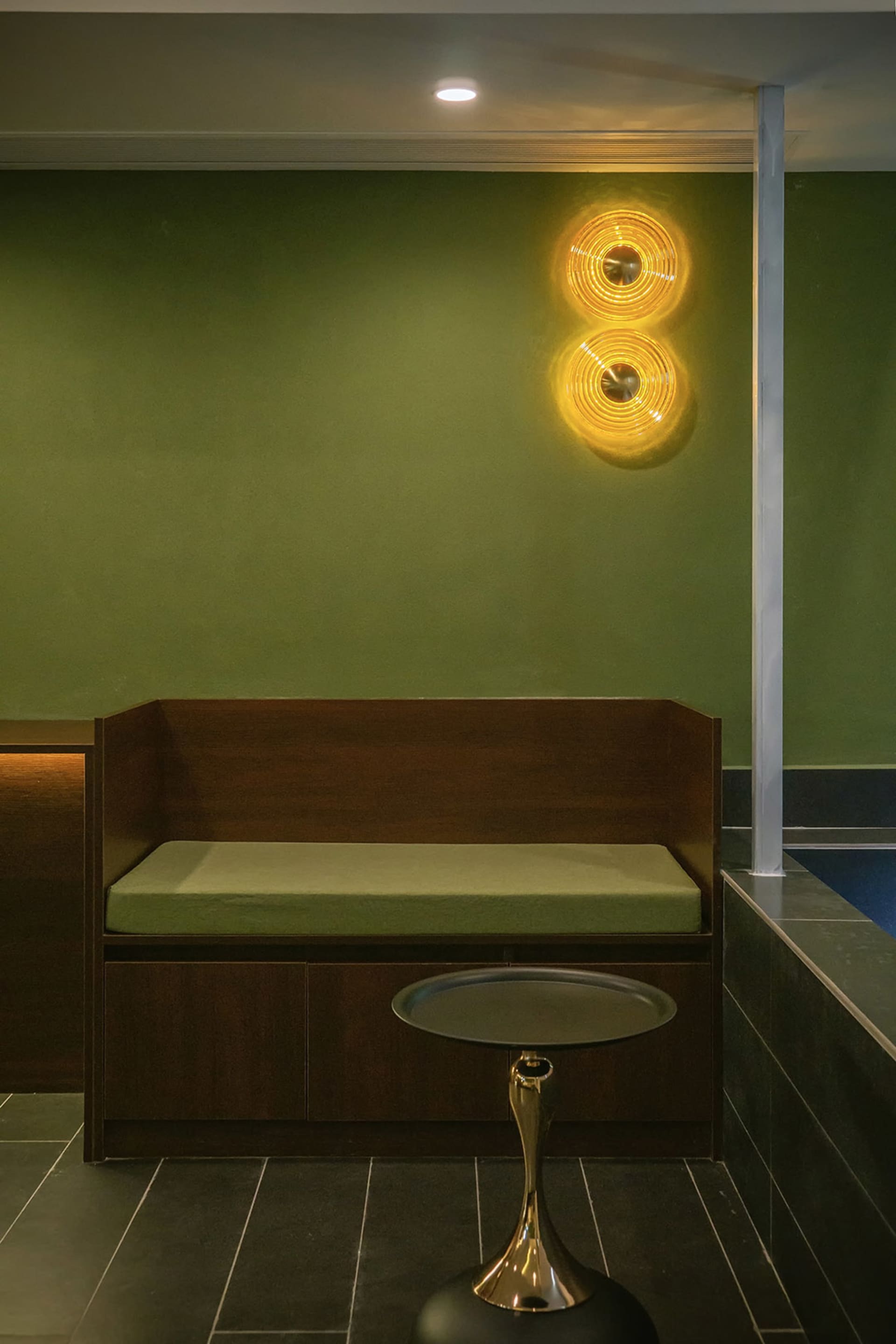 A small, cozy room with green walls, a wooden bench, and a green cushion, lit by a wall light