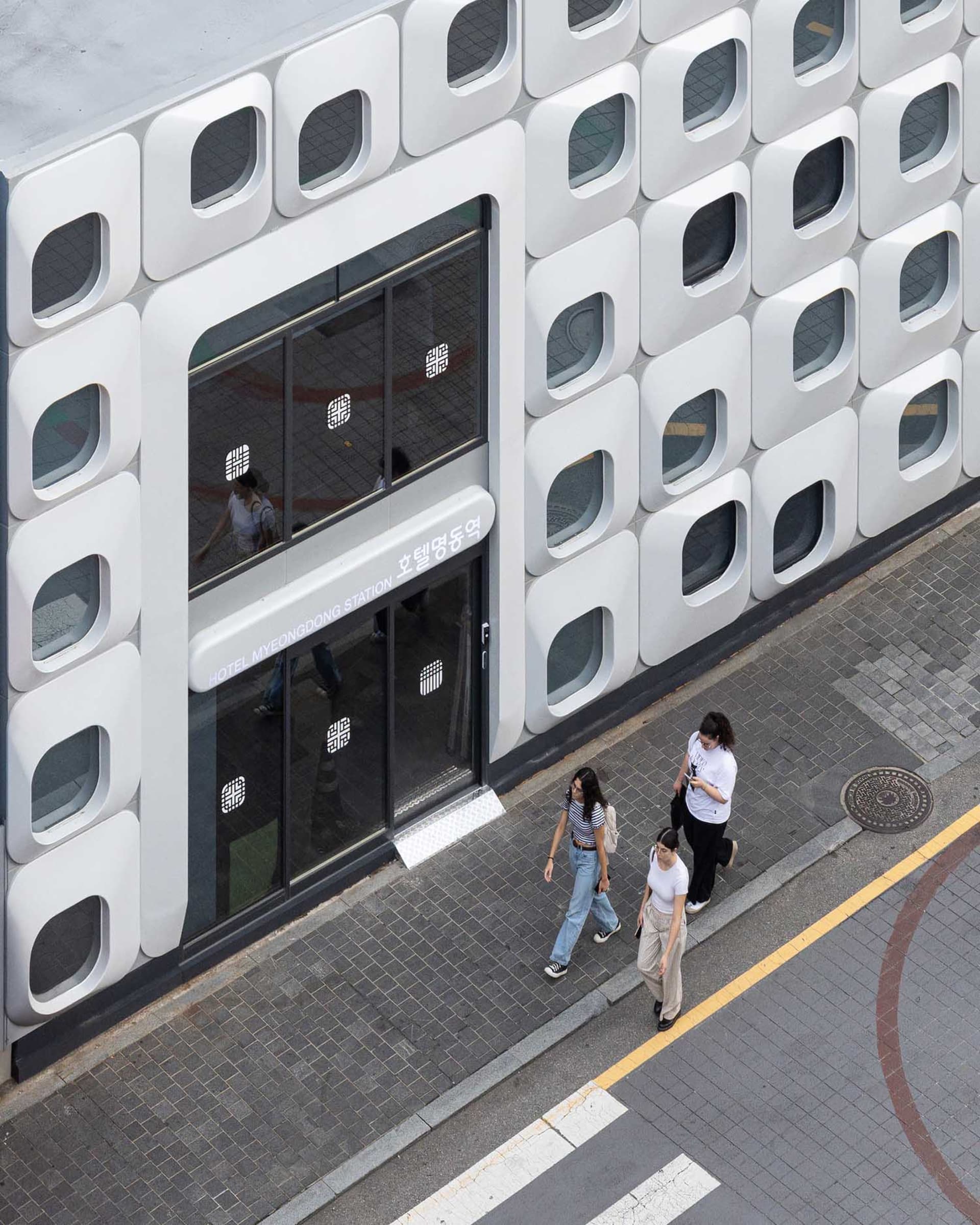 A group of people walking on a brick sidewalk in front of a modern building with a white facade and large windows, set in an urban environment