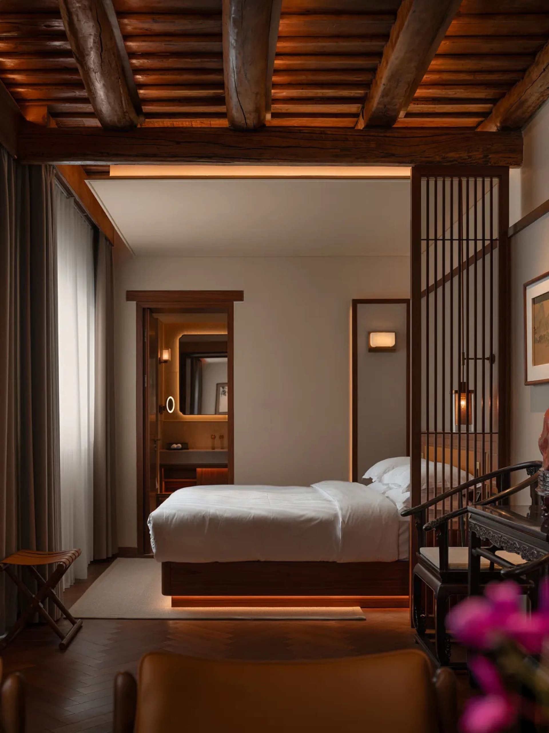 A hotel bedroom with a bed, chair, and desk, featuring wooden furniture and dim lighting, creating a cozy atmosphere
