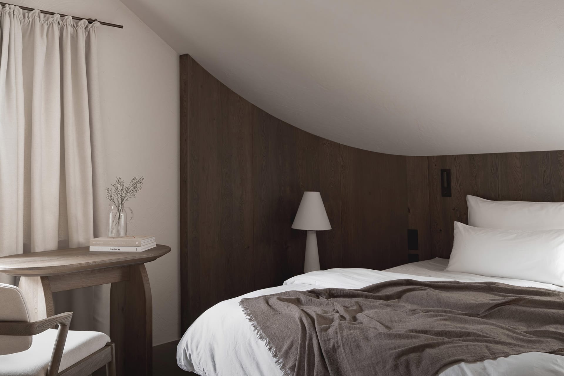 Cozy bedroom with a wooden bed frame, a wooden floor, and a dimly lit atmosphere, featuring a bed, a table, and a lamp