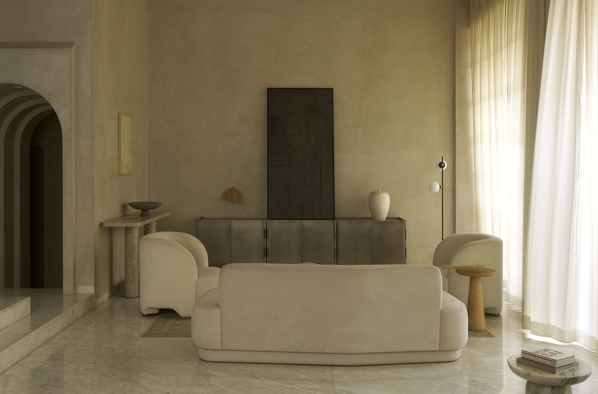 A cozy living room with a beige color scheme, featuring a large couch, a chair, and various decorative pieces, with natural light streaming in through the windows