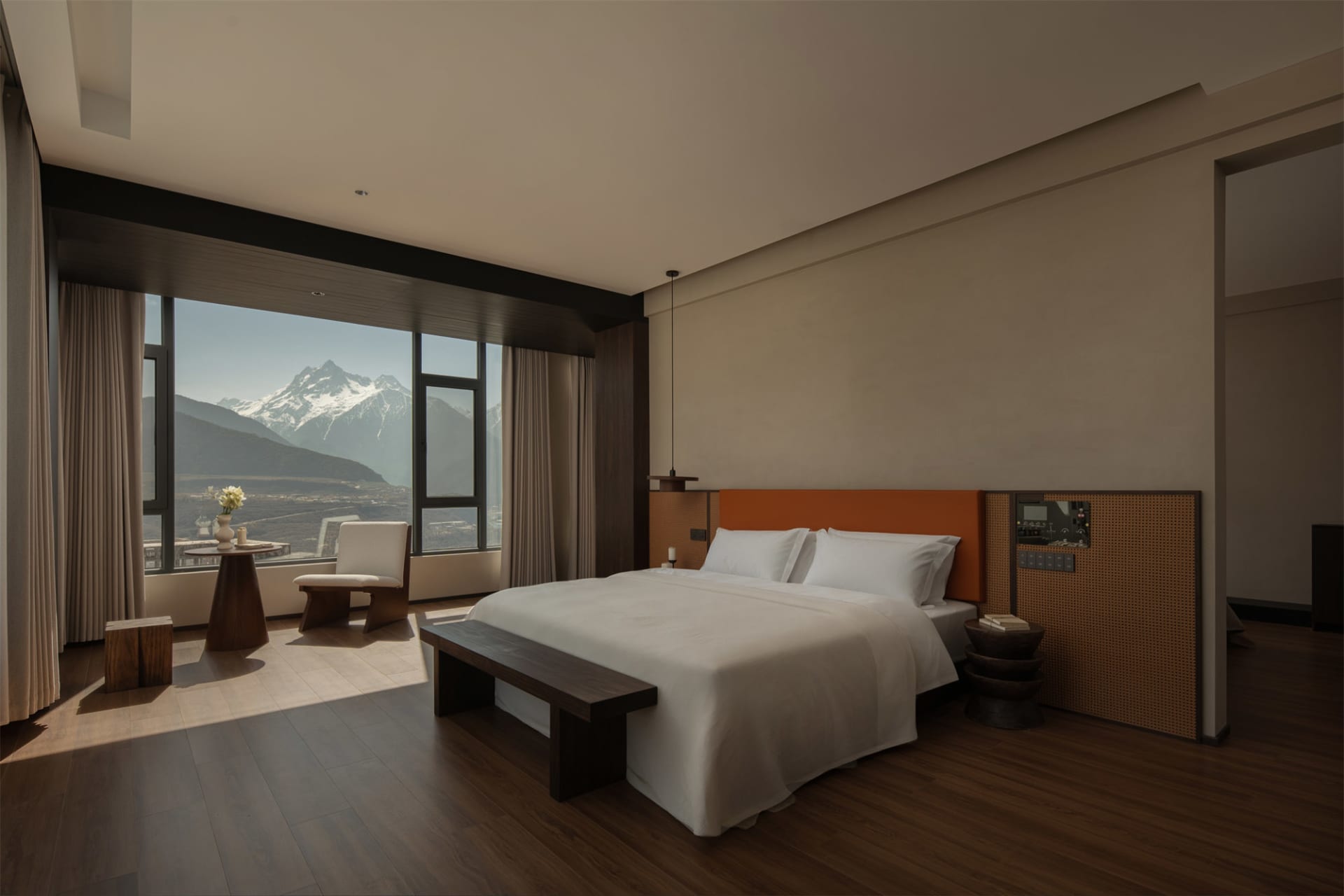 A cozy bedroom with a bed, chair, and table, featuring a large window with a view of the mountains, and a warm atmosphere created by natural light