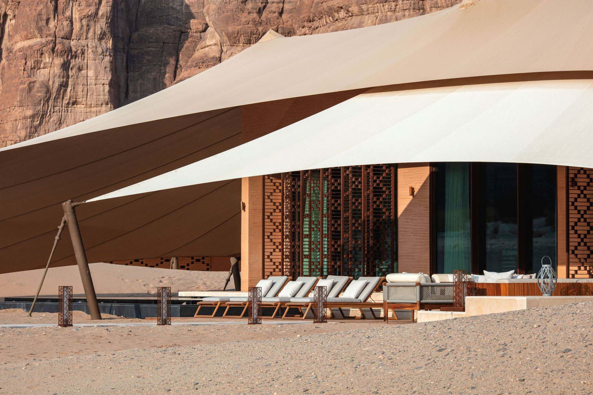 A house with a unique wooden frame structure and a patio area with a dining table and chairs, set against a sandy beach background