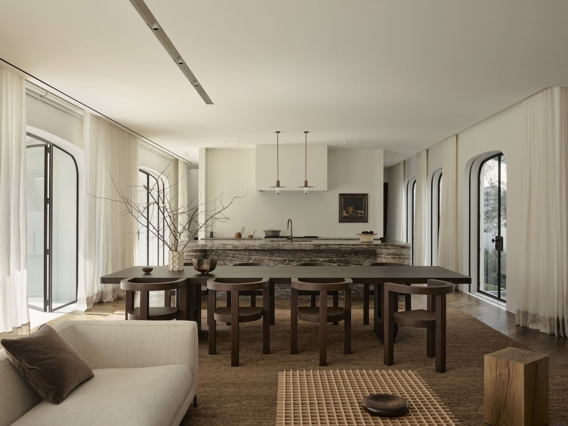 A warm and inviting kitchen and dining area with a large dining table, kitchen counter, and couch, decorated with a vase, bowls, and a potted plant, with natural light streaming in through the windows