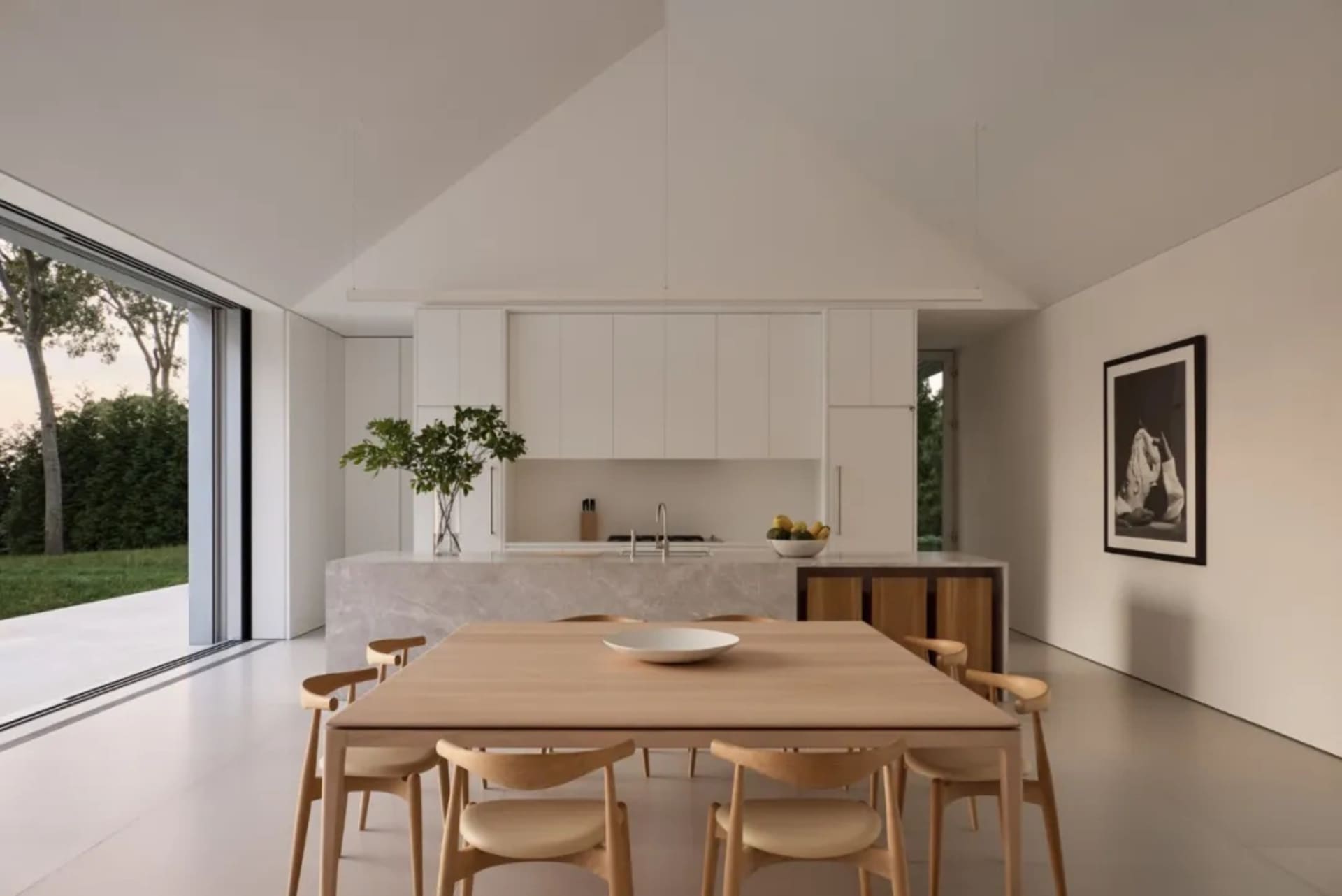 A modern kitchen with a wooden dining table, white cabinets, and natural light from a window, set with a bowl and oranges