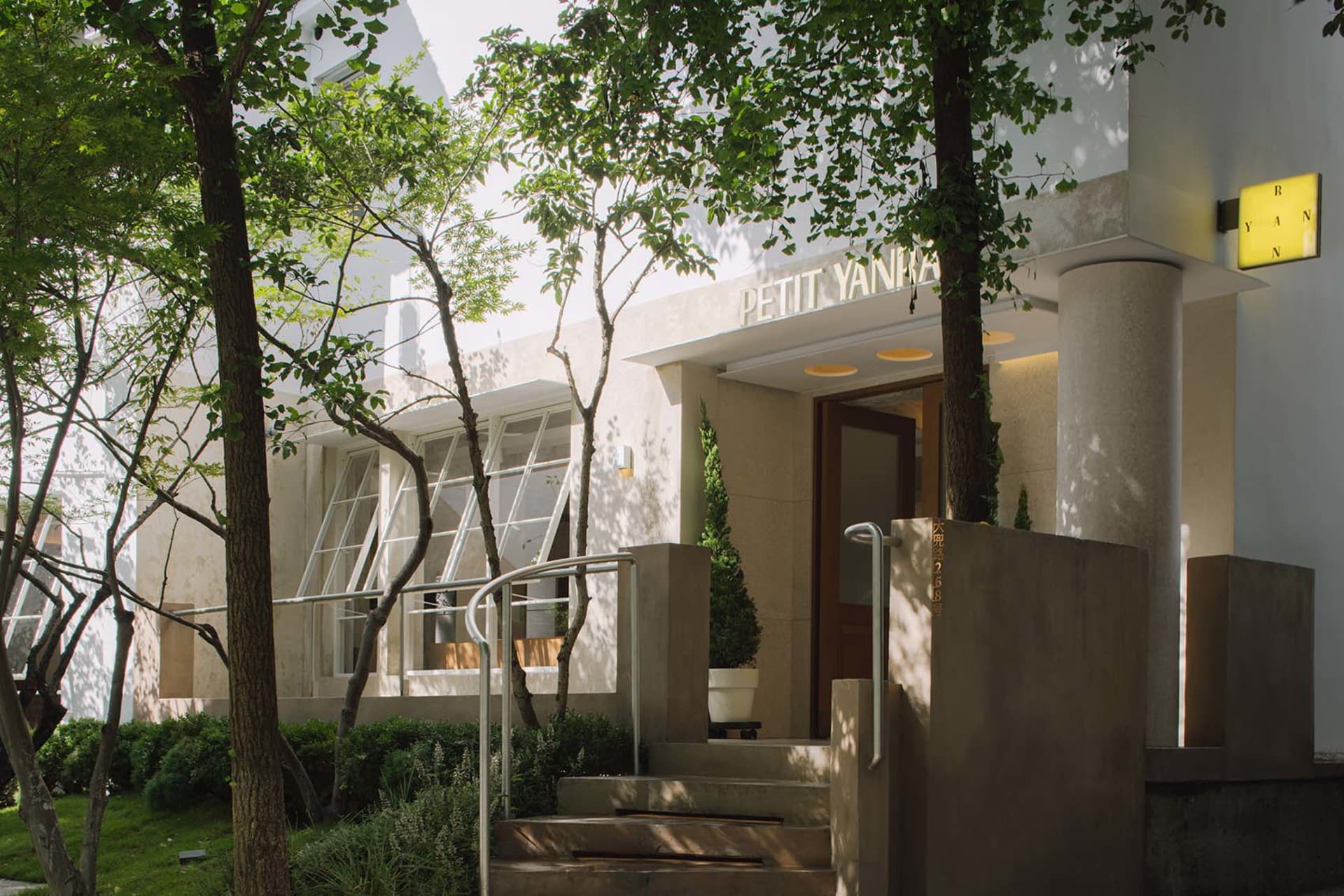 A well-lit exterior view of a white building with a wooden staircase and metal railing, leading to an open doorway revealing a hallway