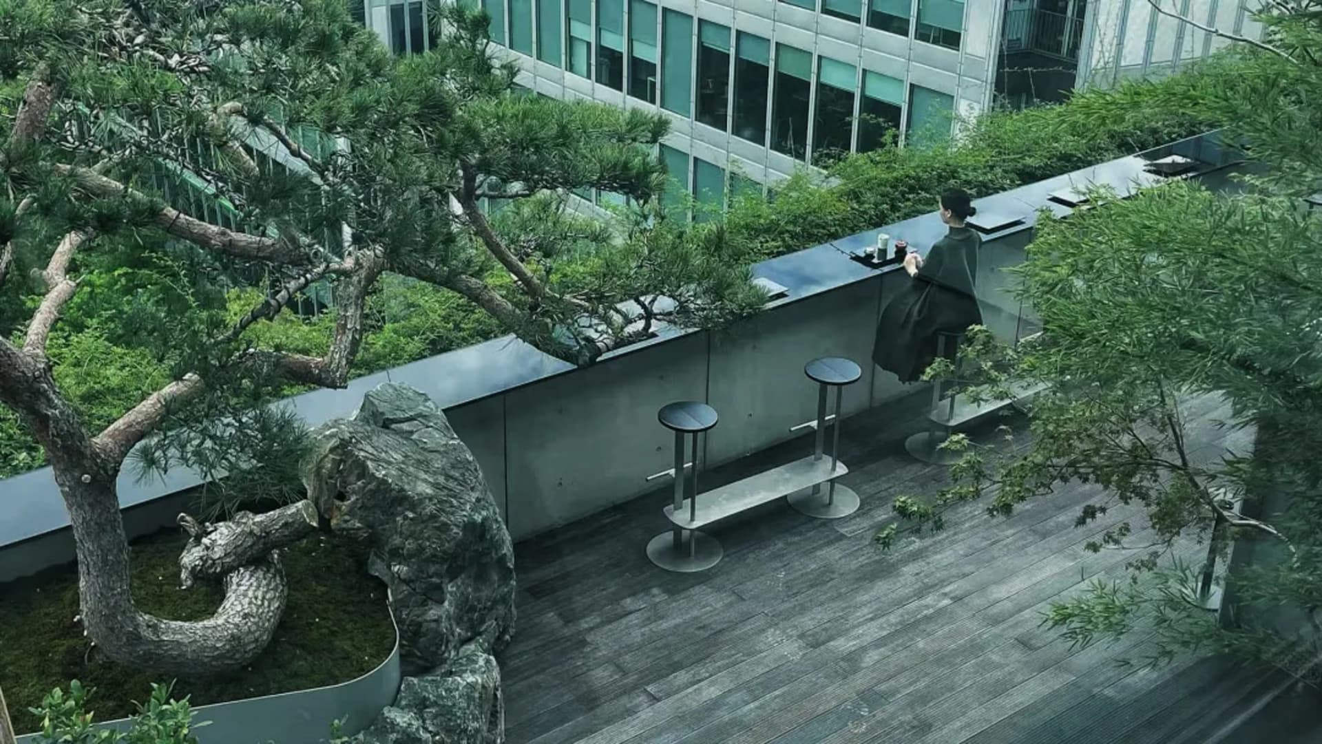 A woman sits on a bench in a modern rooftop patio with a table and chairs, surrounded by a potted plant, set in a relaxed and inviting urban environment