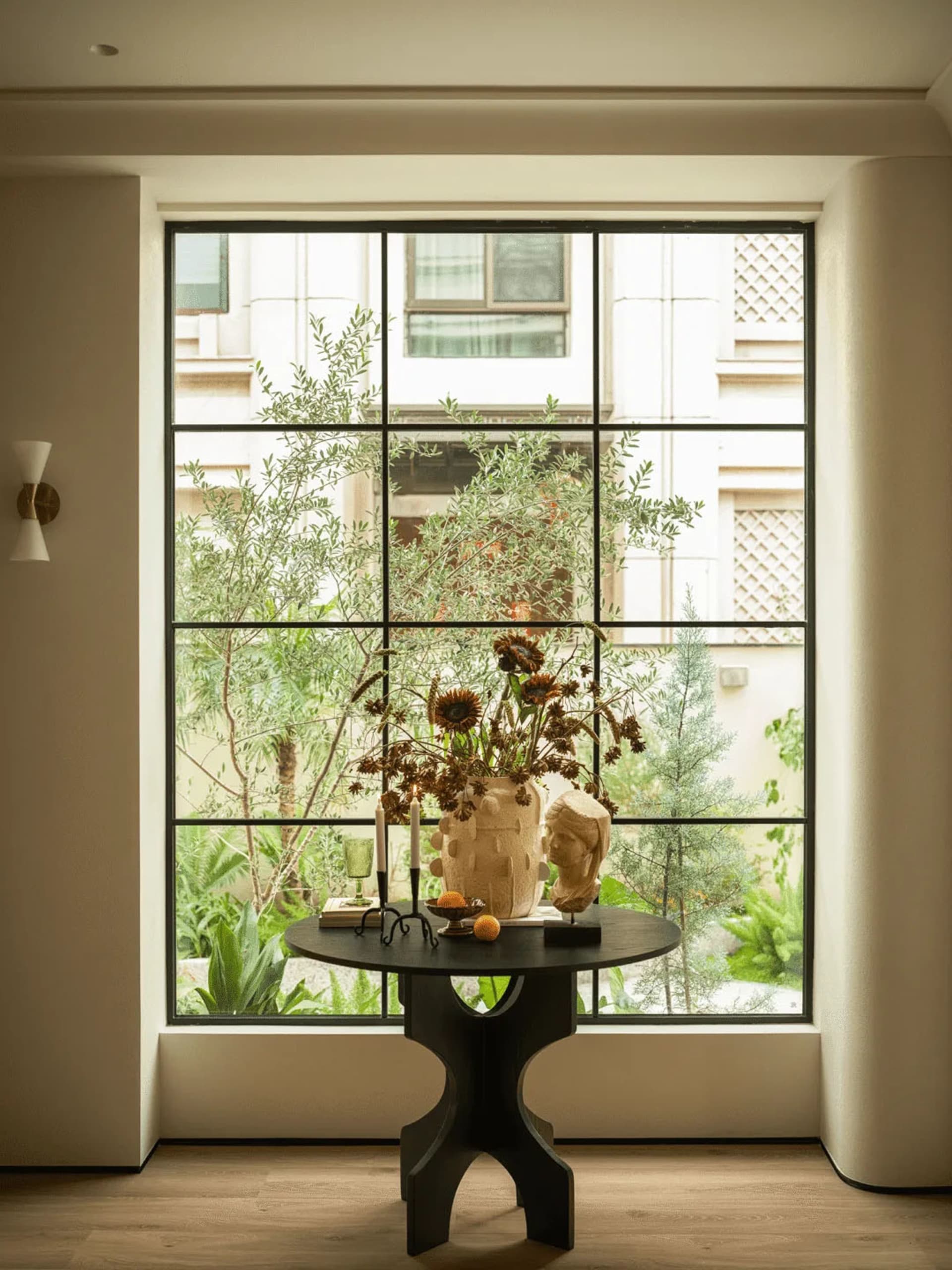 A living room with a table in front of a large window, featuring a vase of flowers and a candle, surrounded by natural light