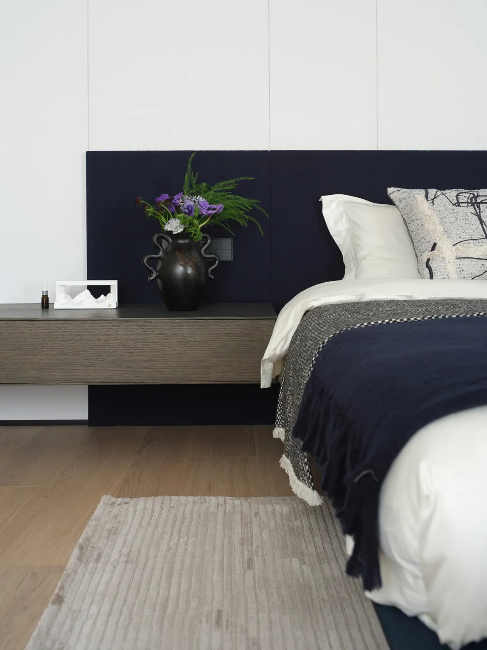 A well-lit bedroom with a bed, nightstand, and vase with flowers, featuring predominantly white materials and a clean appearance