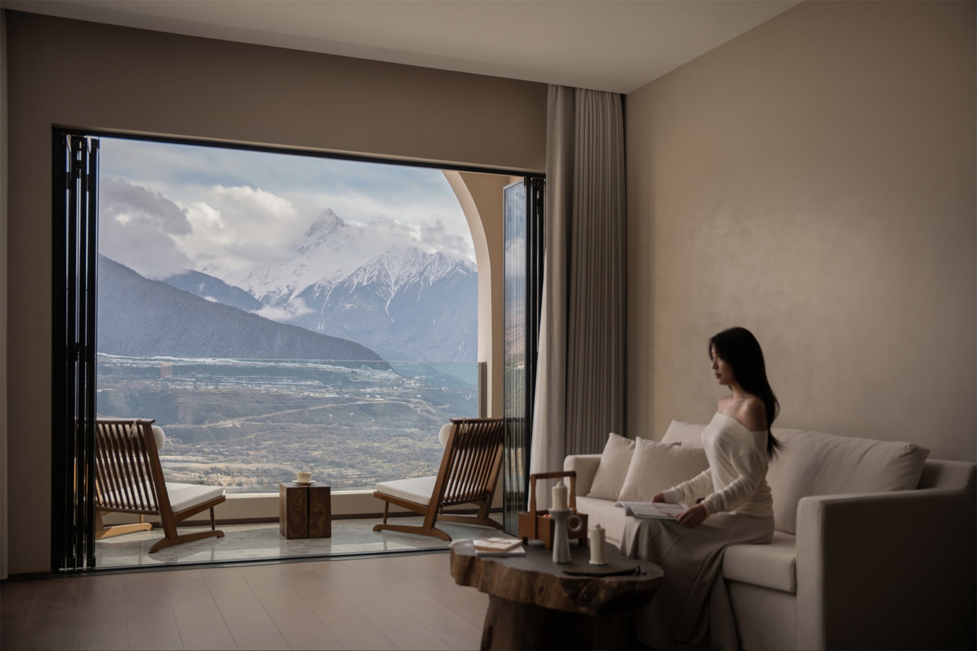 Serene living room with a woman enjoying a mountain view from a large window, surrounded by a couch, chair, dining table, and potted plant