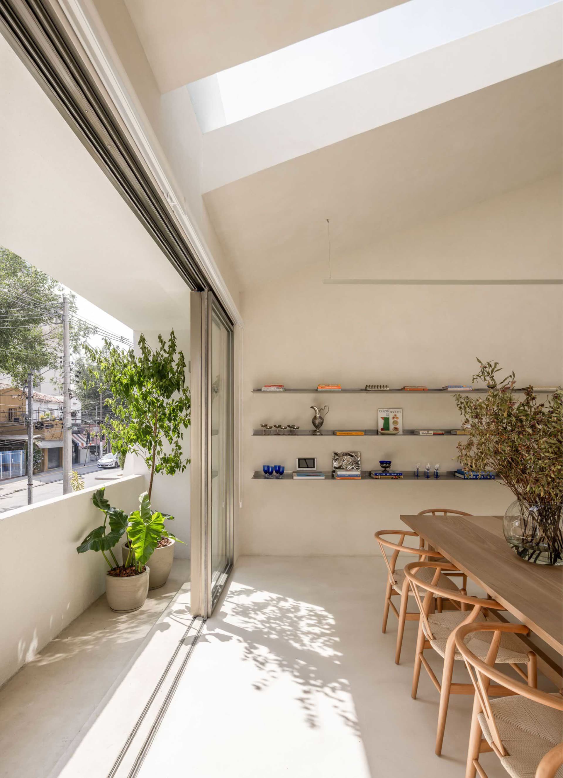 A modern living-dining space with a large window, glass door, and skylight, featuring a dining table, chairs, and a potted plant, with predominantly white materials and natural light