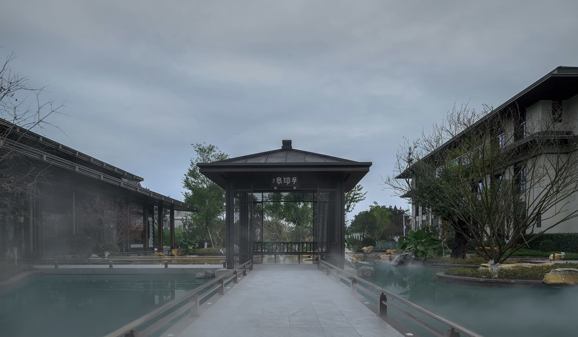 一張照片，展示著有一個黑色屋頂的建築物，環繞著樹木，展示著一座木橋跨越水池，通往一座亭台