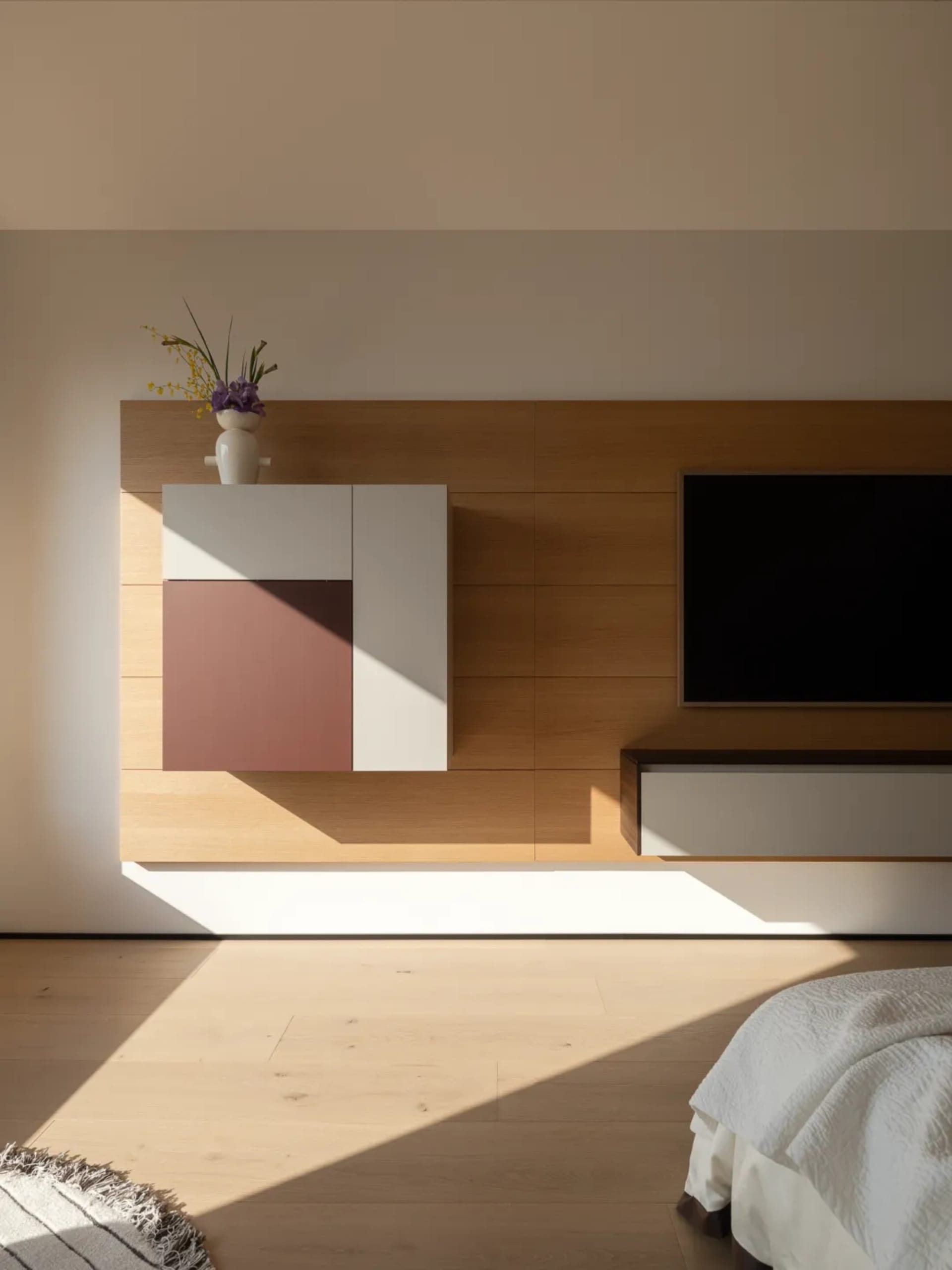A modern living room with a wooden floor and white walls, featuring a flat-screen TV, cabinet, and vase with flowers, with natural light and a clean aesthetic