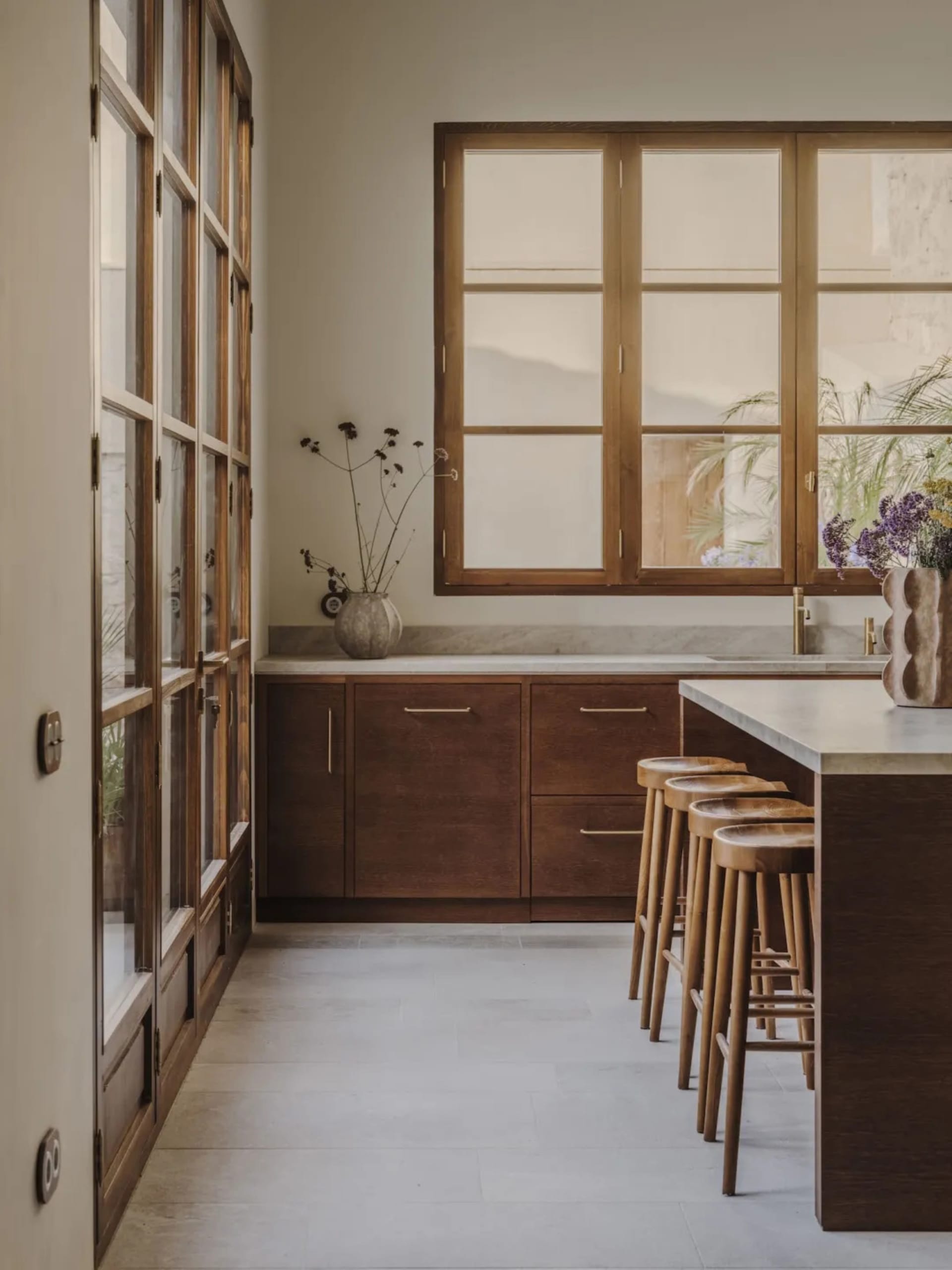 A well-lit kitchen with a wooden floor, marble countertop, sink, refrigerator, and stools, with a window providing natural light