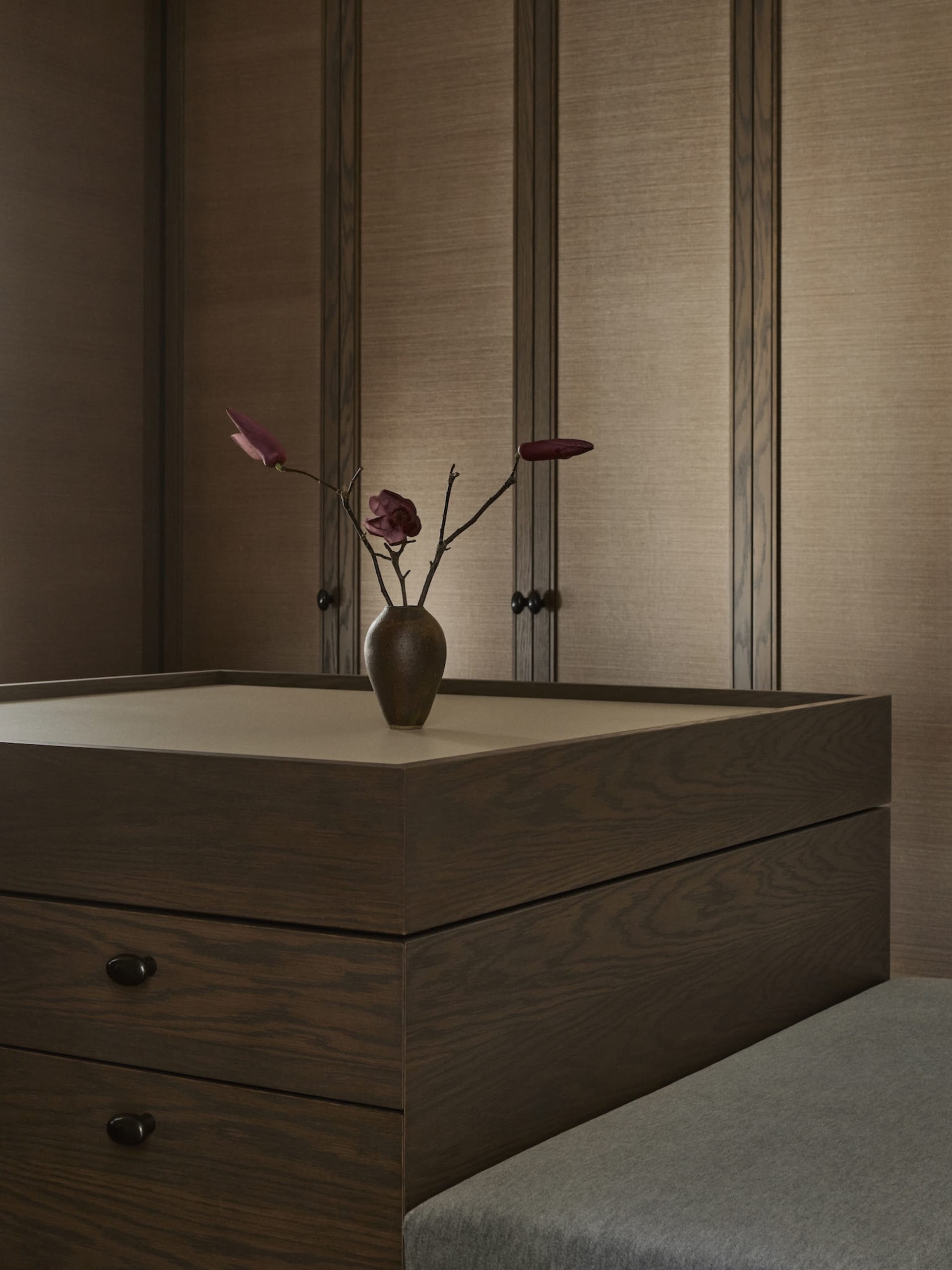 A cozy bedroom with a wooden dresser, a vase of flowers, and a bed in the background, creating a warm and inviting atmosphere