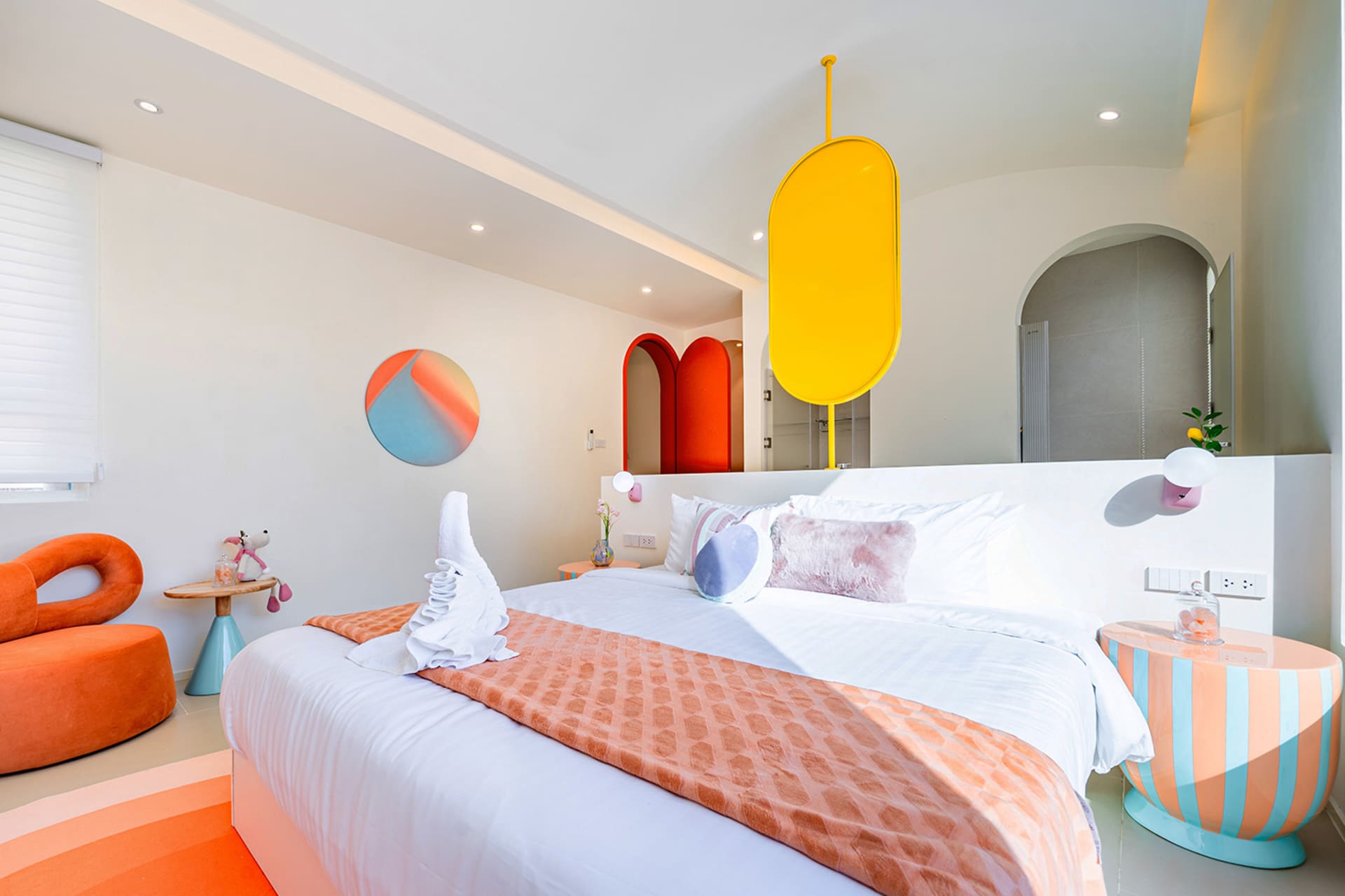 Cozy bedroom with a bed, chair, and yellow and orange color scheme, featuring a white sheet and red and orange checkered blanket, with natural light streaming in through a window