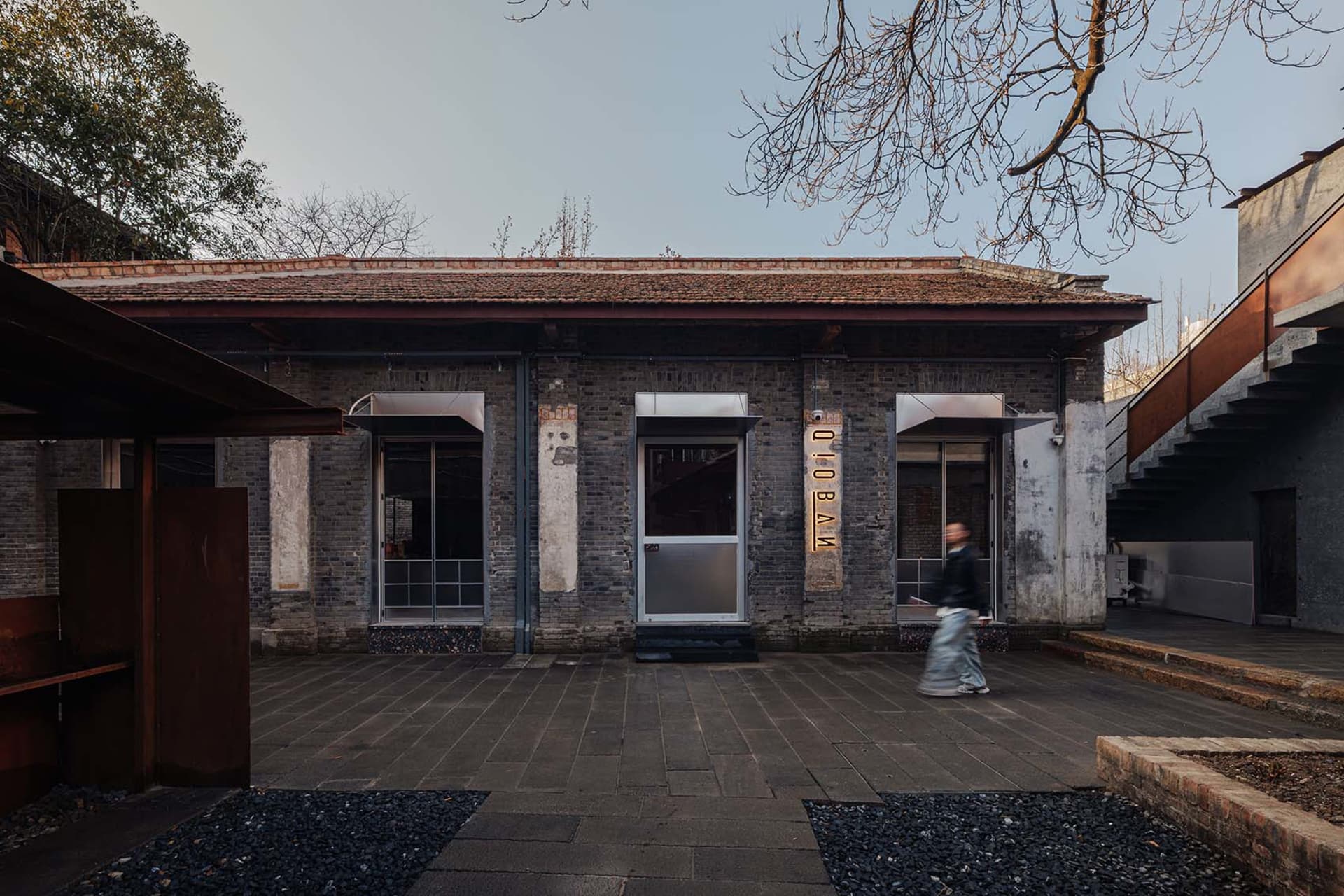 A woman walks towards a brick building with a tiled roof, set in an outdoor scene