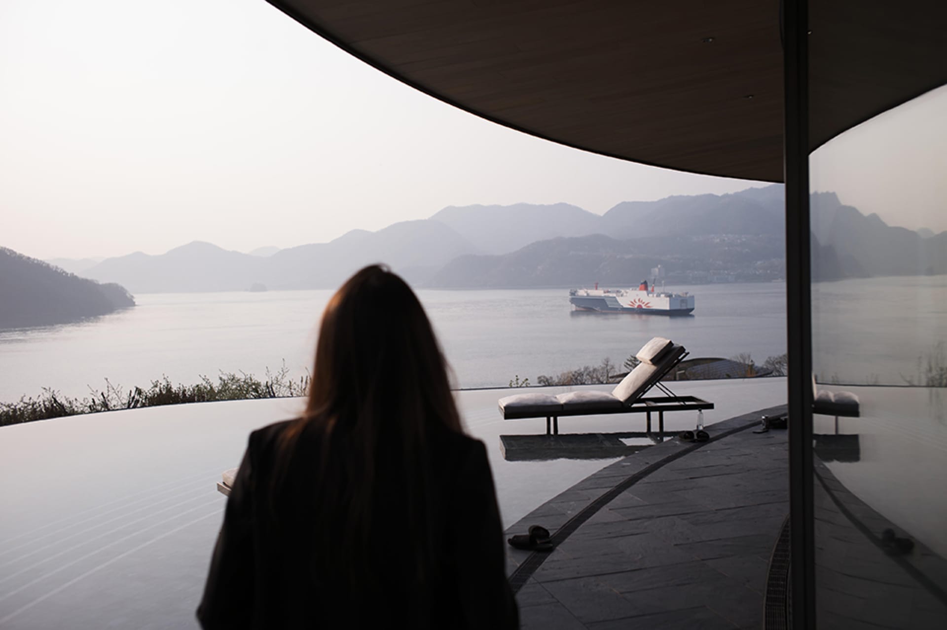 A woman stands on a glass balcony overlooking a large body of water, with a boat visible in the distance, set in a modern upscale home