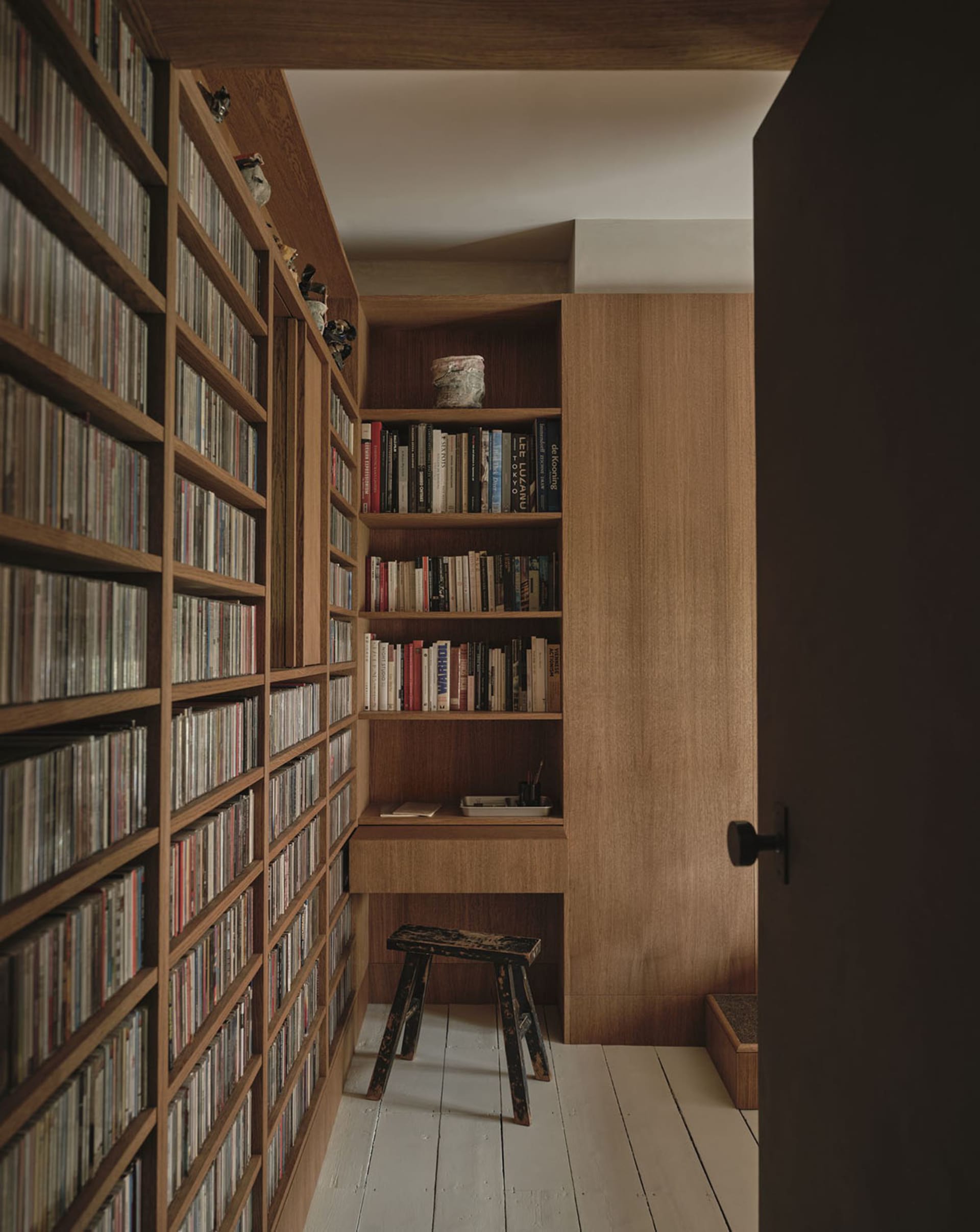 A cozy room with a wooden bookshelf in the corner, a chair in front, and books of various sizes neatly arranged on the shelf, creating a warm and inviting atmosphere
